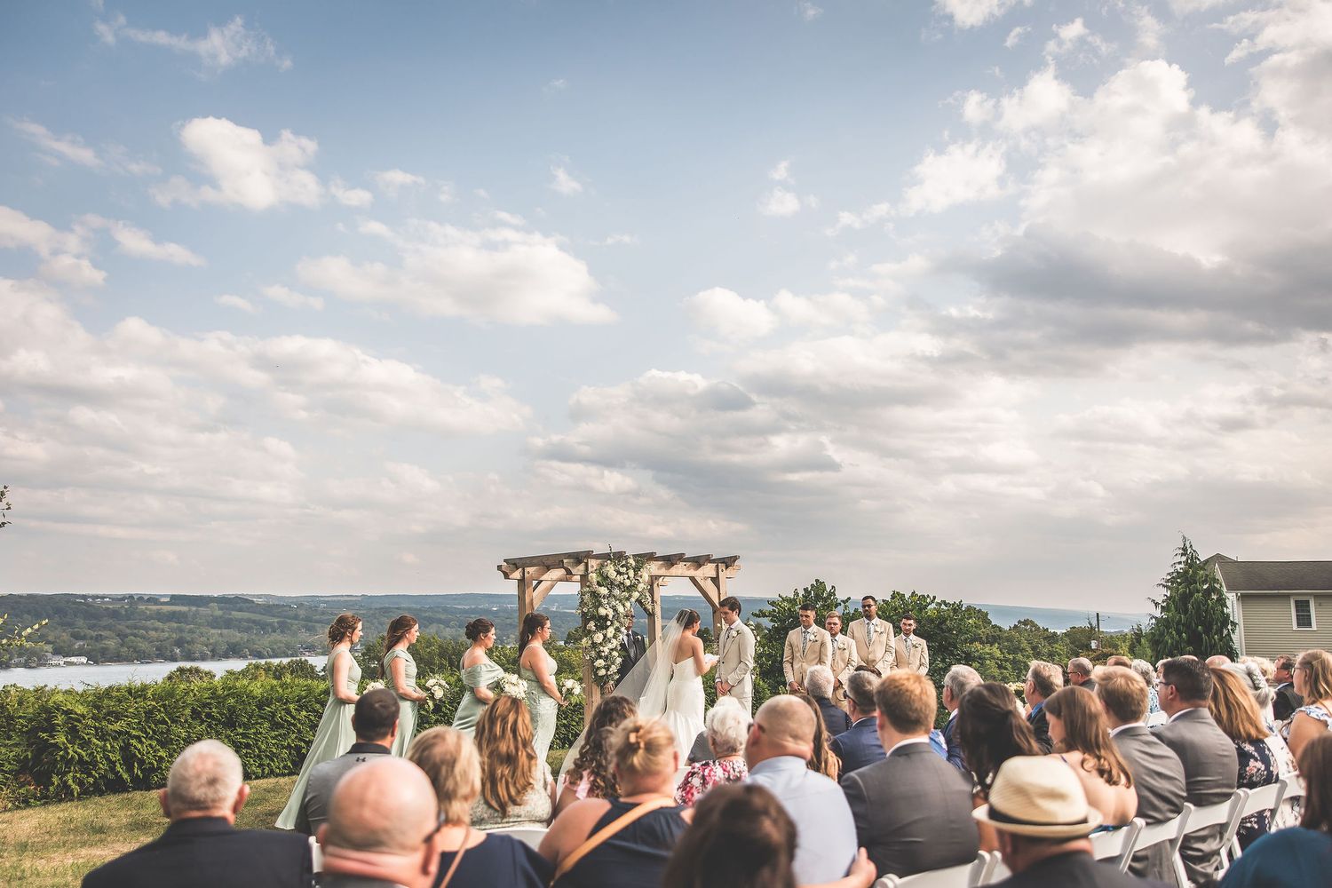 Scenic hilltop wedding ceremony with wooden arch and white chairs overlooking water.