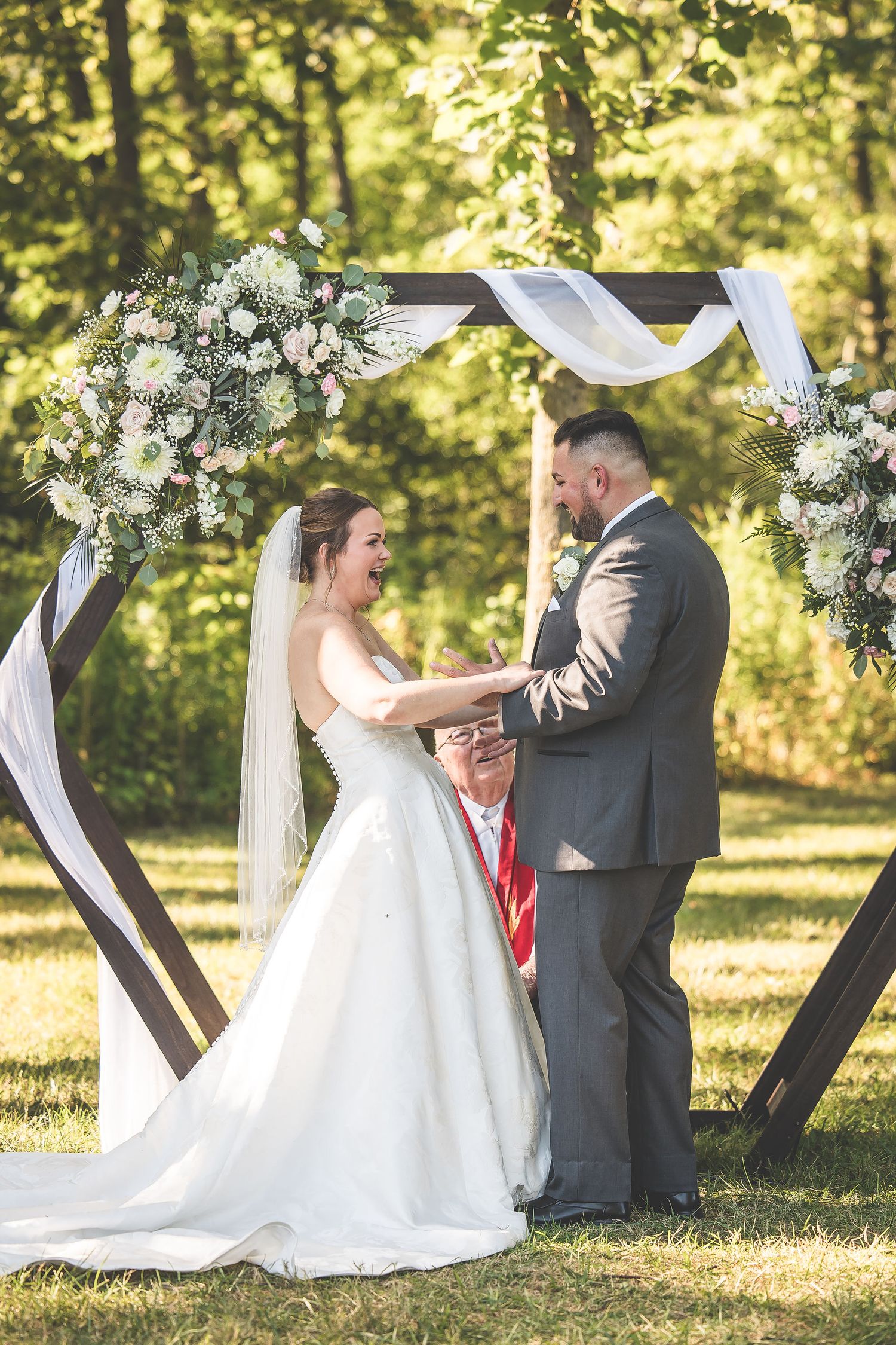 A joyful wedding ceremony moment captured under a floral-adorned arch in natural sunlight.