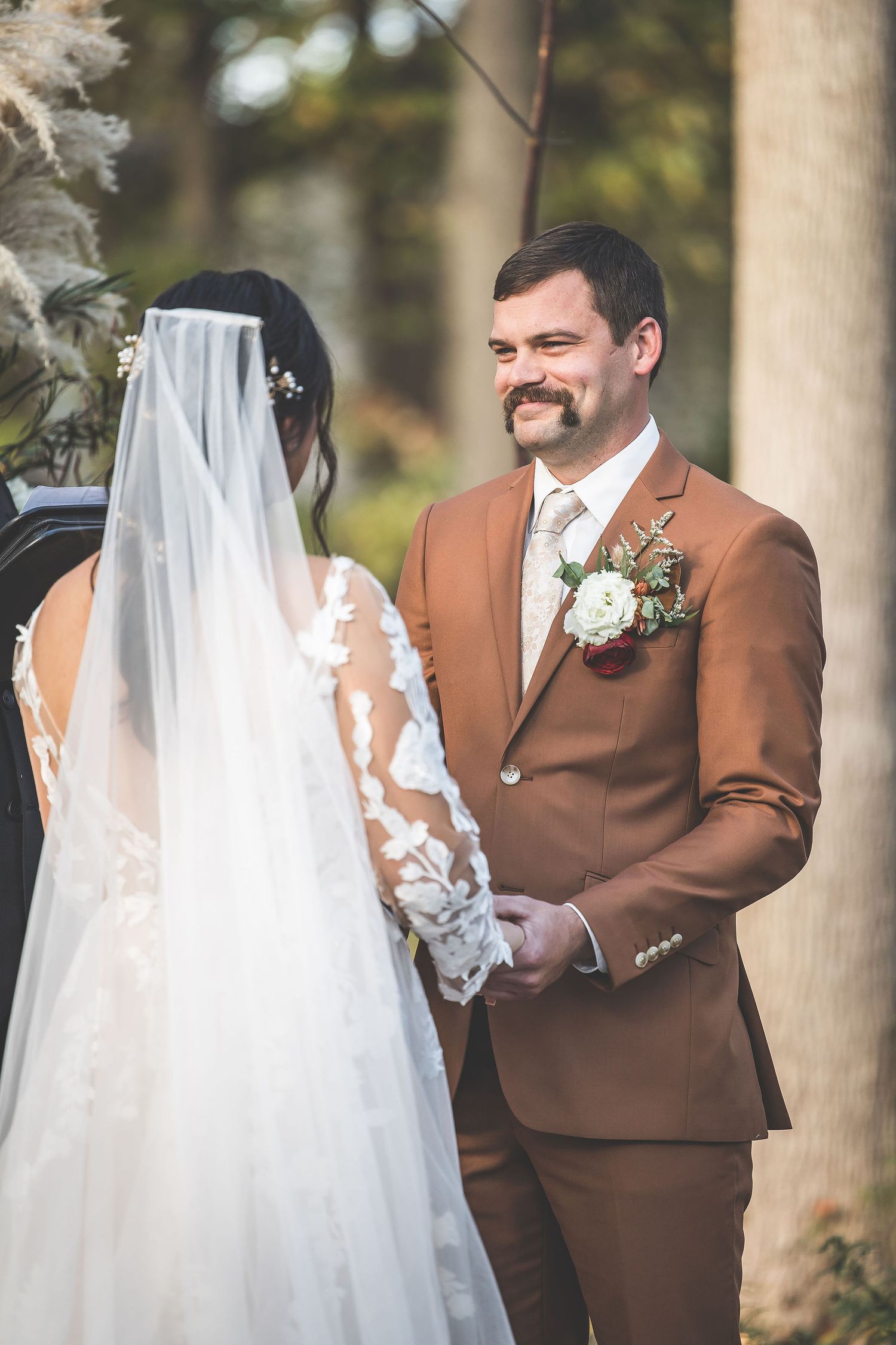 Wedding couple exchanging vows under tall trees with bride wearing detailed lace gown.