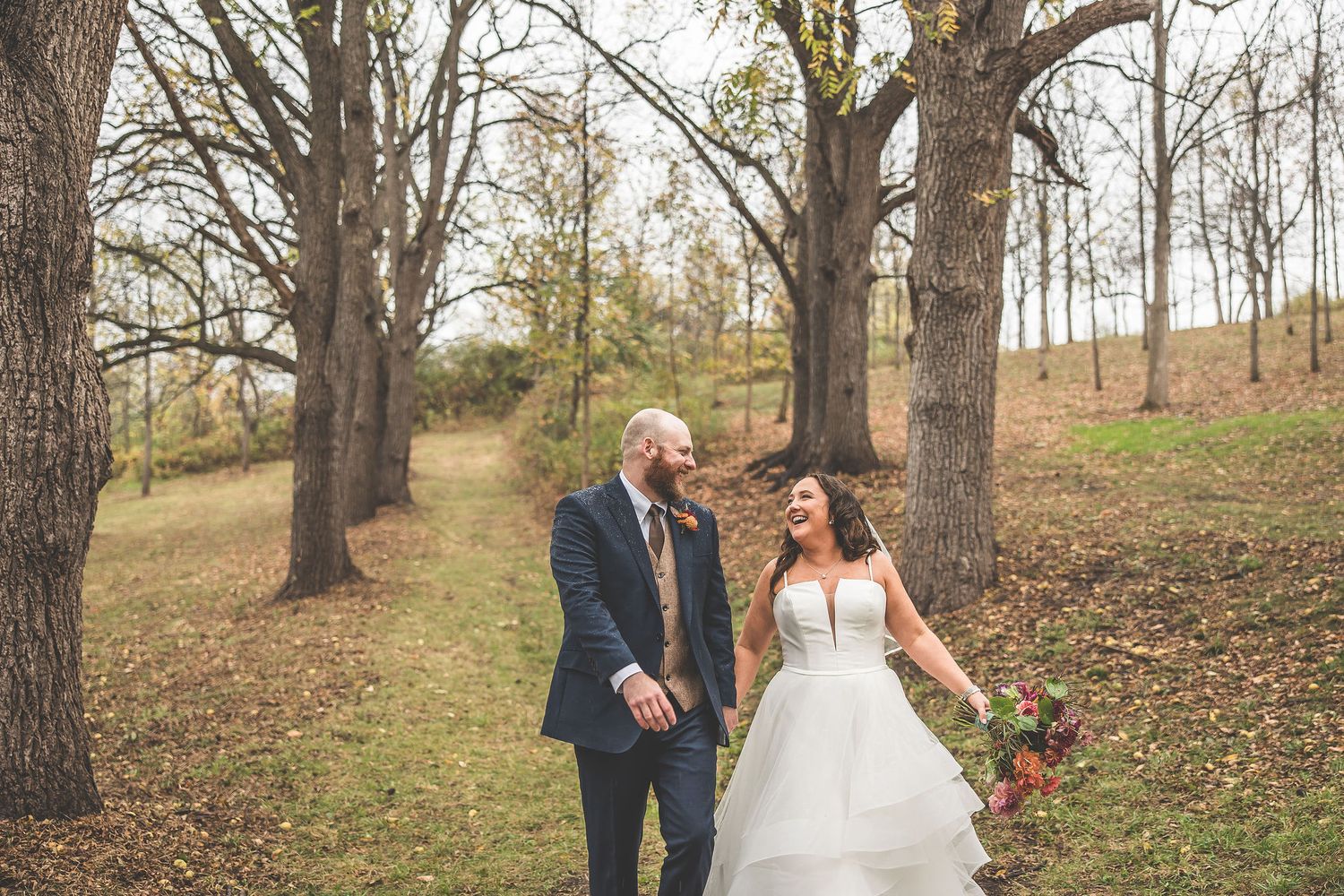 A romantic autumn wedding photo of a couple walking together through a wooded path with fallen leaves.