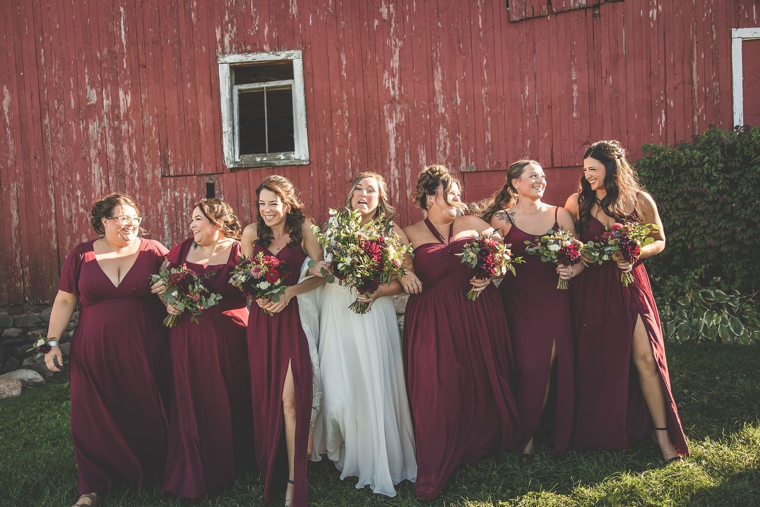 Wedding party poses in front of rustic red barn with bridesmaids in burgundy dresses and white bridal gown.