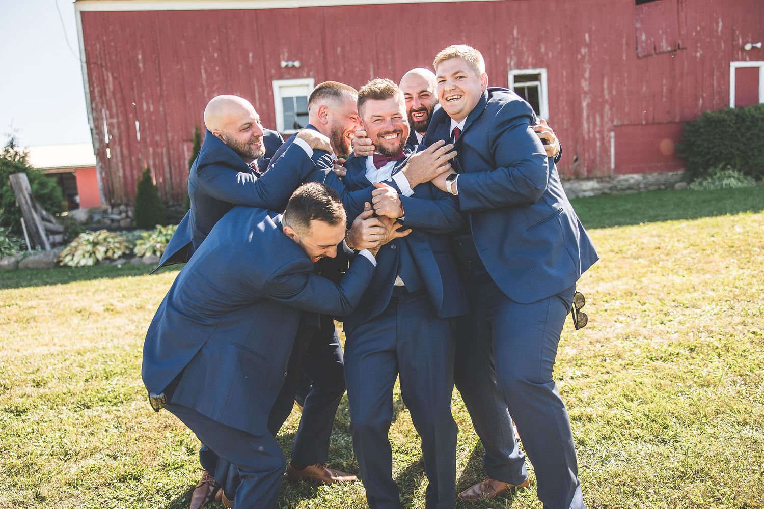 Groomsmen in navy suits share candid laughing moments outside red barn.