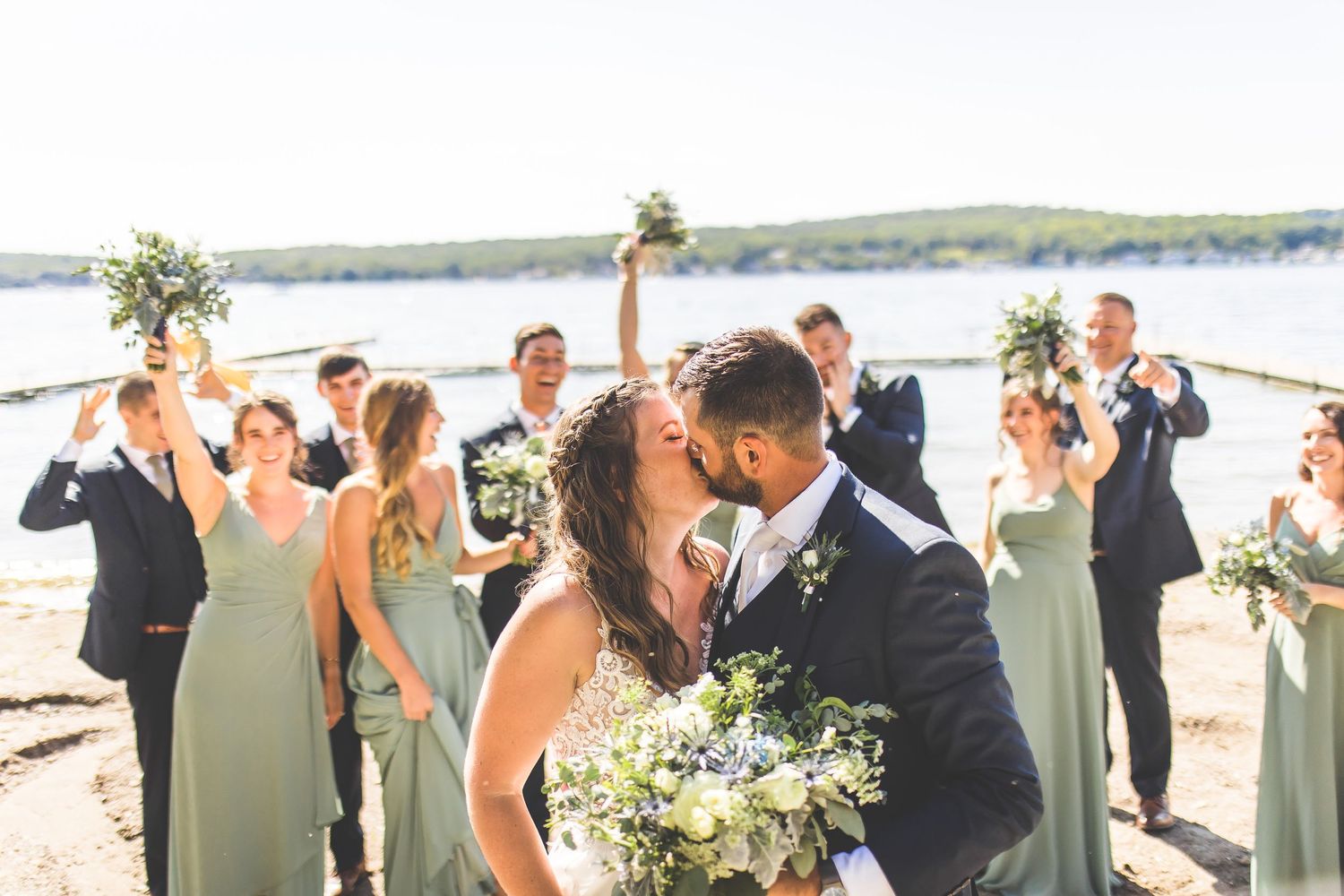 Wedding couple shares romantic moment surrounded by wedding party in sage green dresses.