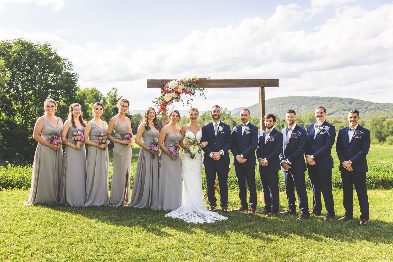 Large wedding party in gray dresses and navy suits poses on grassy hillside.