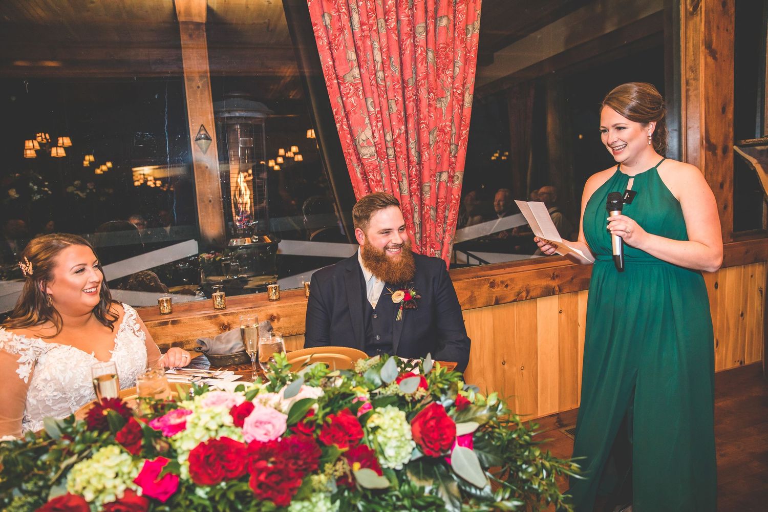 Wedding party seated at decorated tables with vibrant red flower centerpieces.