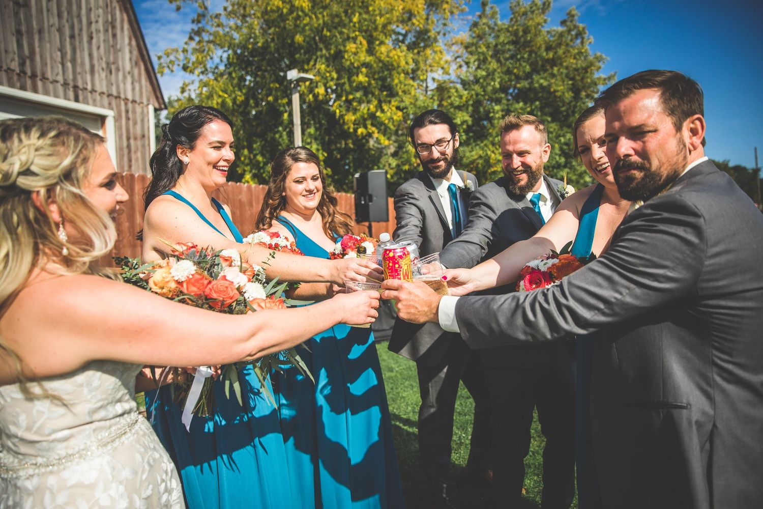 Wedding party toasts with drinks outside a rustic barn on a sunny day.