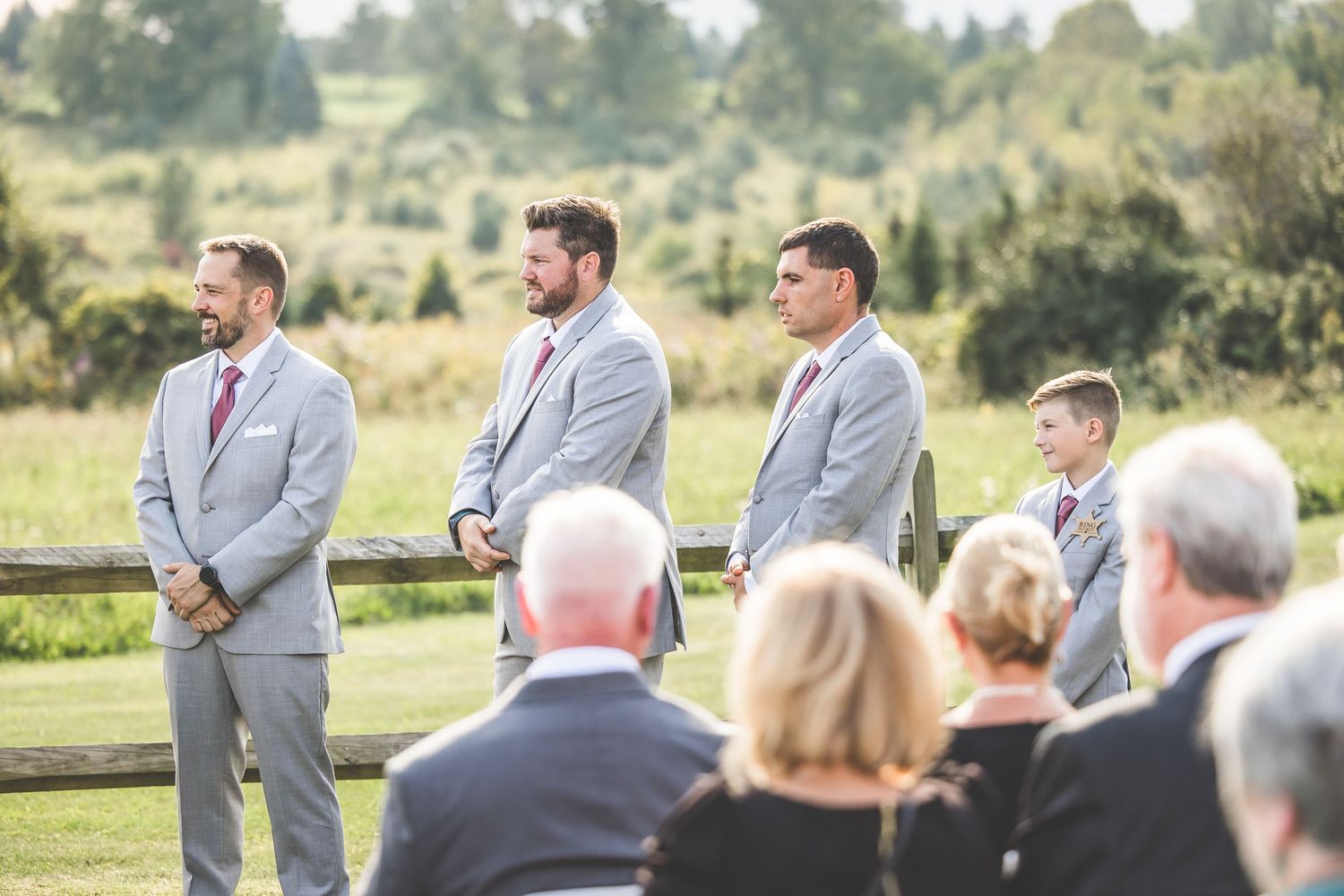 Groomsmen in grey suits standing at outdoor wedding ceremony with natural landscape.