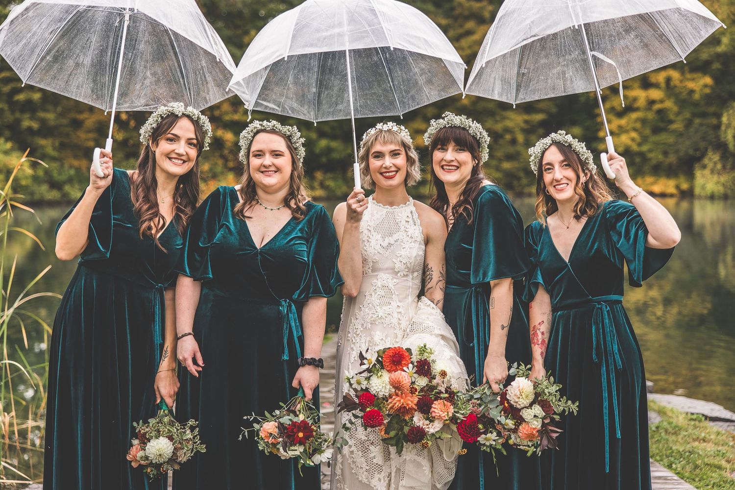 Bridal party in emerald velvet dresses poses with clear umbrellas by lakeside.