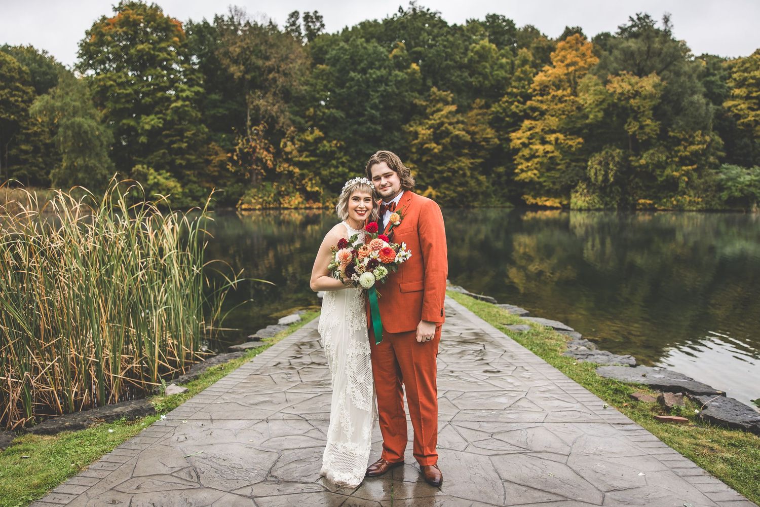 Wedding couple poses on stone path by autumn lake with cattails and colorful fall foliage in background.