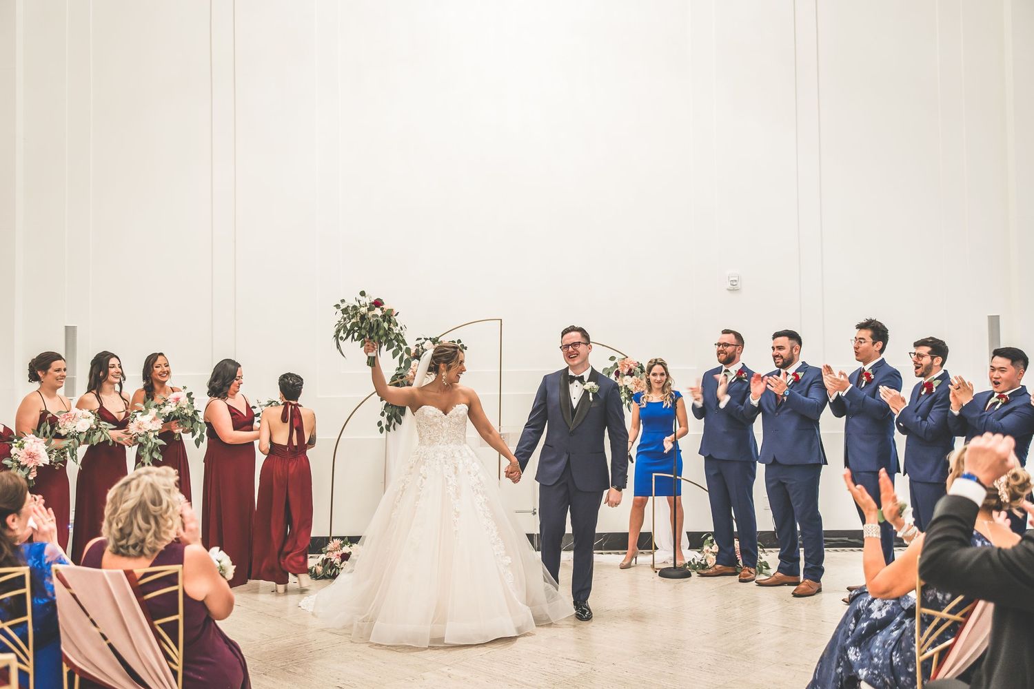 Newlyweds walk down the aisle while wedding party in burgundy and blue attire applauds during indoor ceremony.
