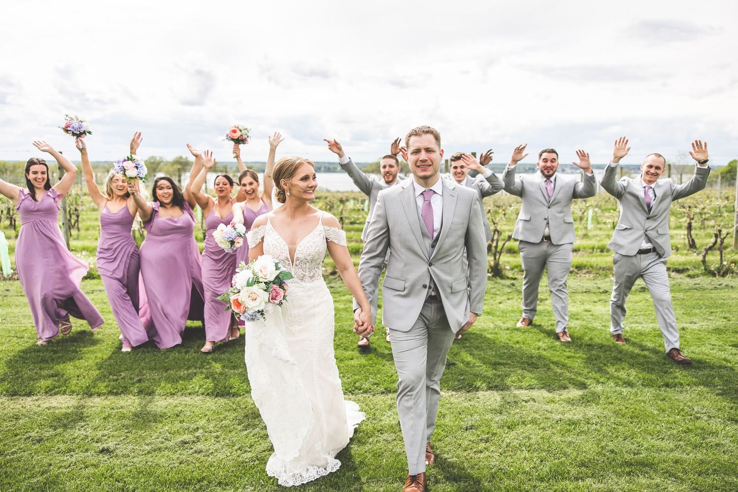Wedding party in purple dresses and gray suits celebrating joyfully in an outdoor vineyard setting.