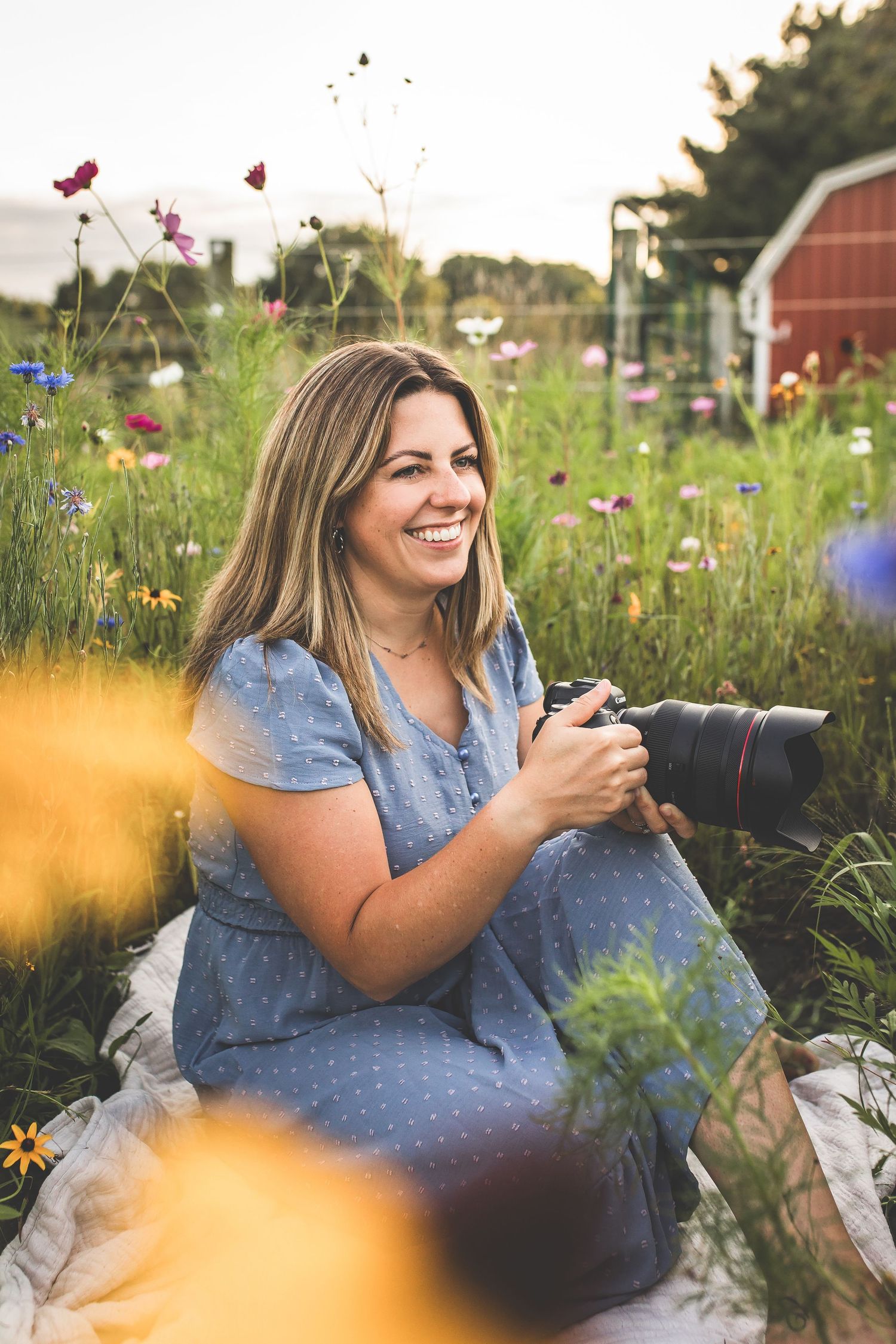 Woman in polka dot dress sits among blooming wildflowers near rustic red barn.