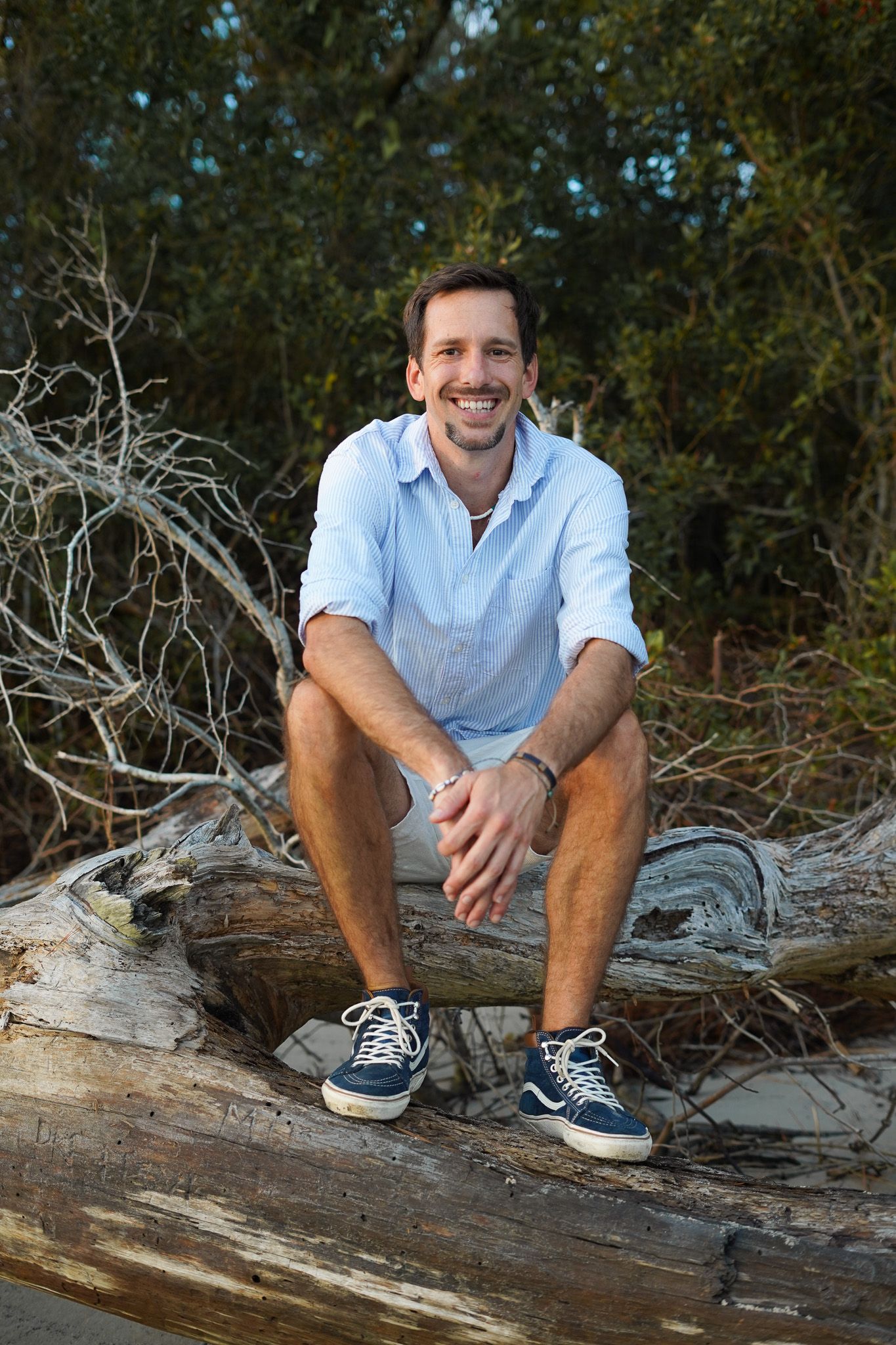 Person in casual attire sits on driftwood at beach wearing blue button-down shirt and shorts.