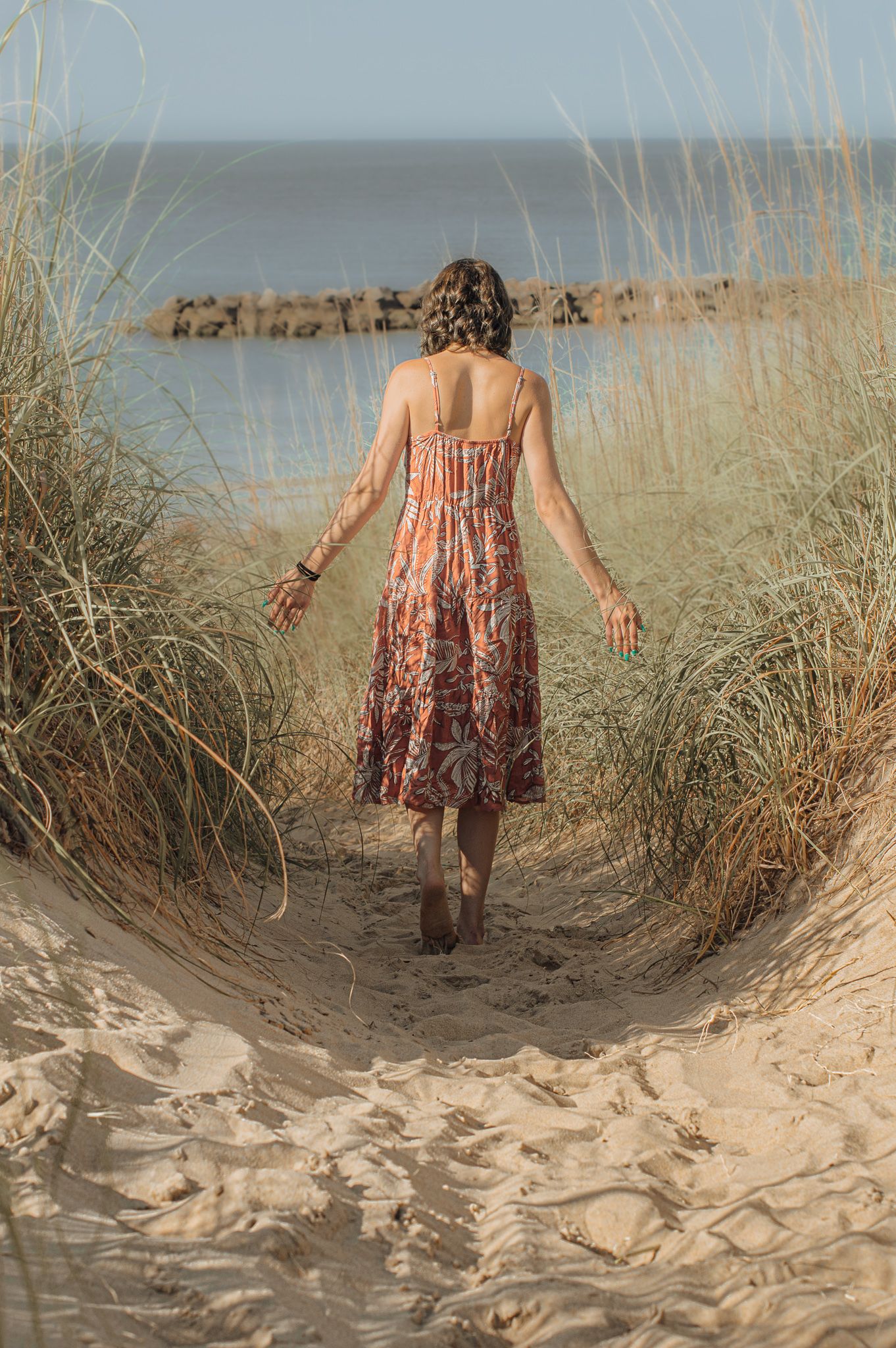 Person in floral dress walks through beach grass dunes towards the ocean.