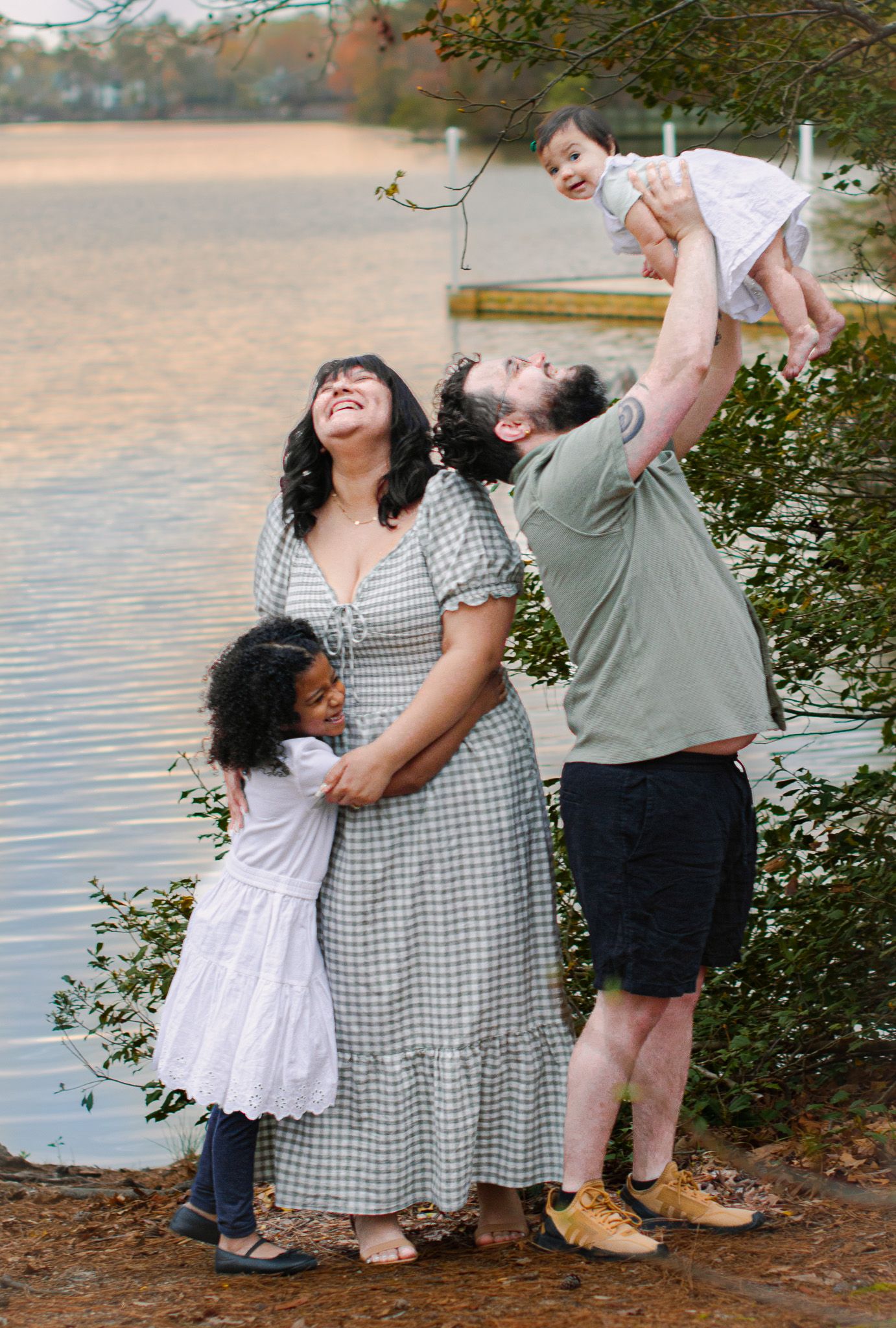 Happy family of four posing by a lake, parents laughing as dad lifts baby in the air while daughter hugs mom.