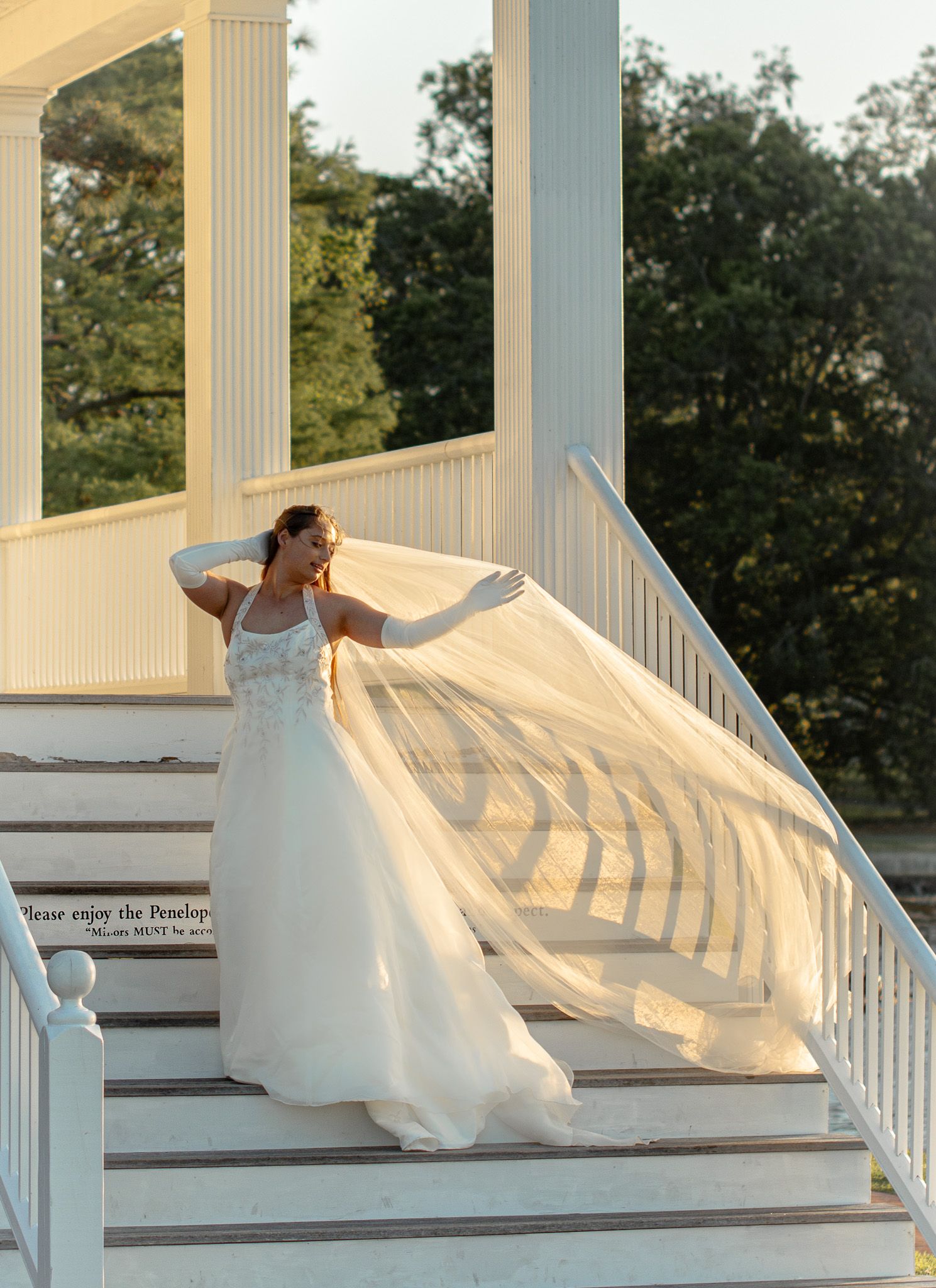 Bride in white gown and veil poses gracefully on white staircase steps at golden hour outdoor venue.
