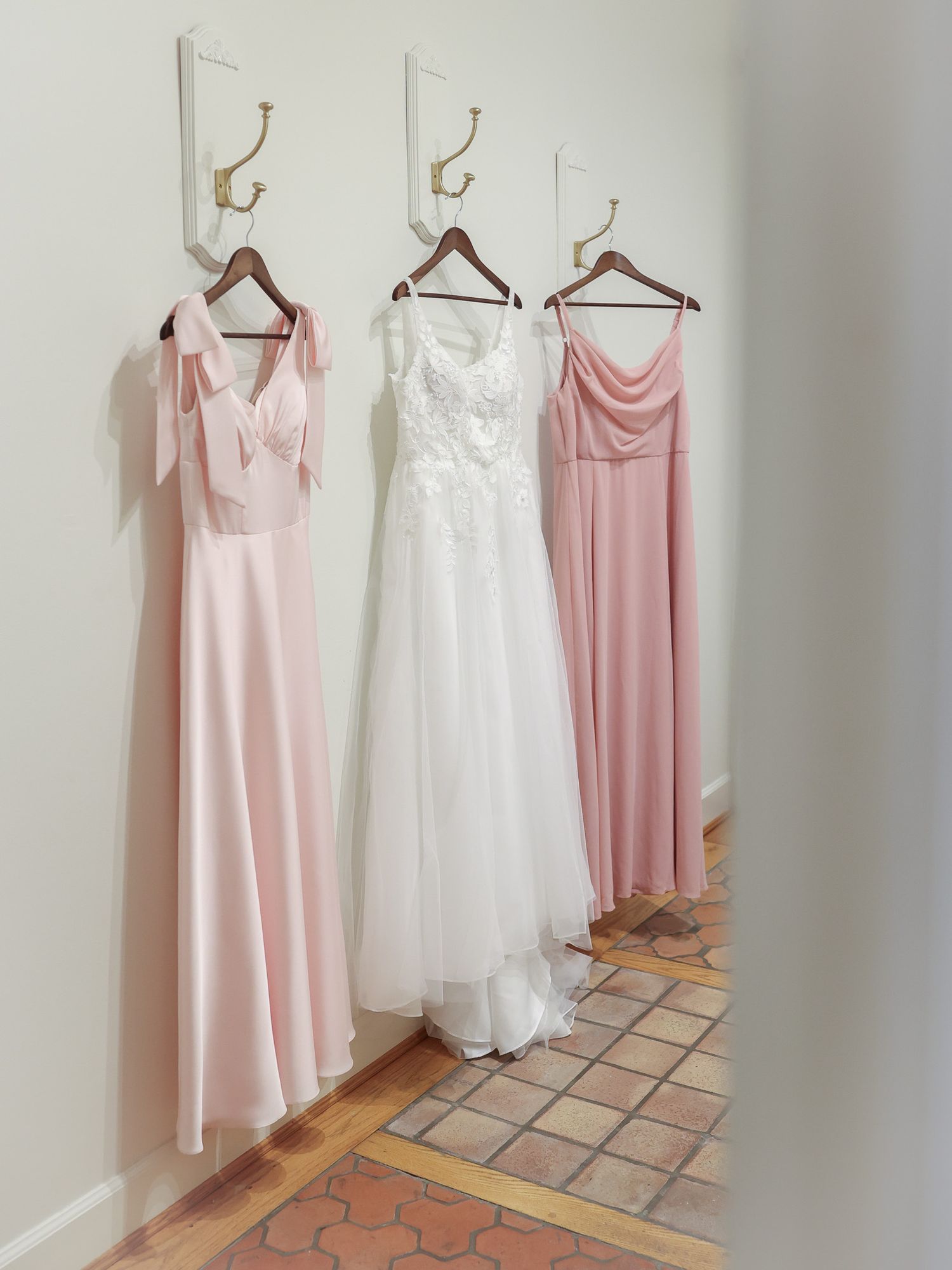 Three dresses hang on wall hooks: two blush pink bridesmaid gowns flank a white lace wedding dress.