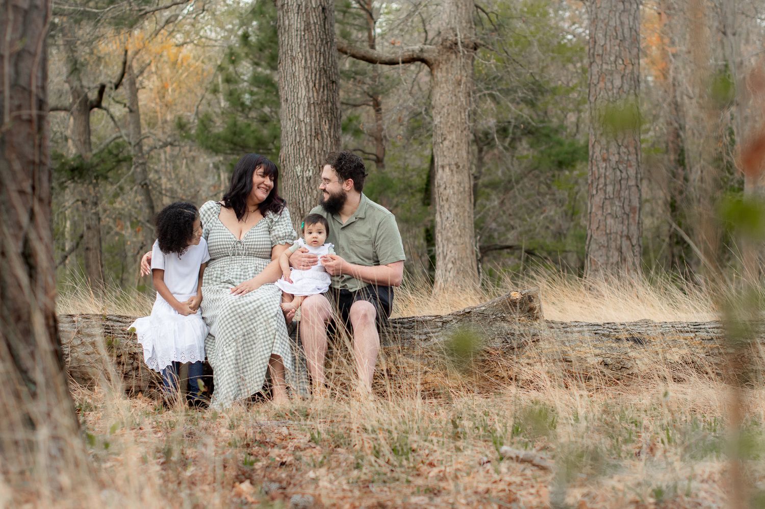 Family of four poses in a wooded forest setting, pregnant mother in green dress with husband and two young daughters.