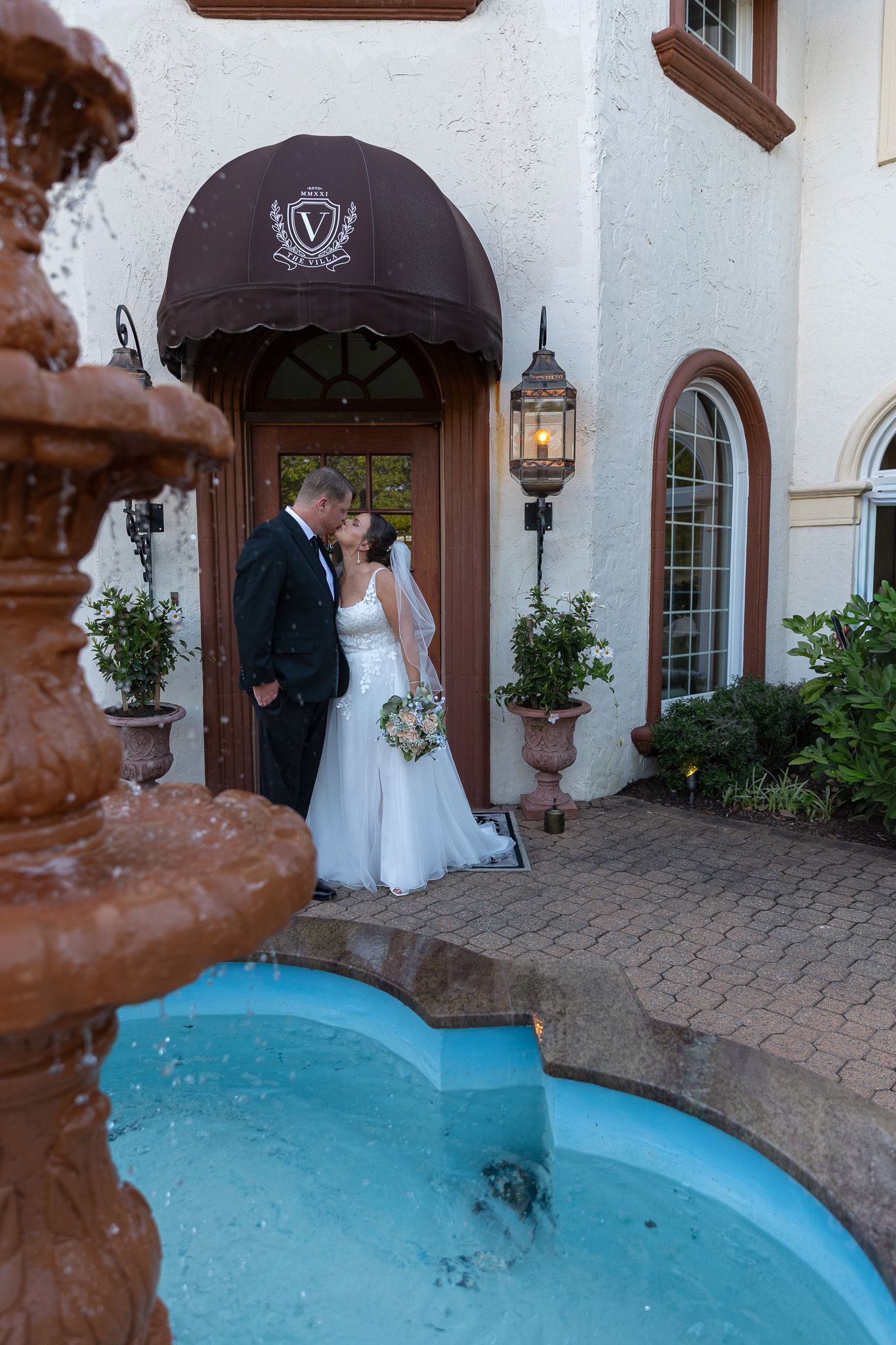 Bride and groom share a kiss near a blue fountain outside an elegant venue with a brown awning.