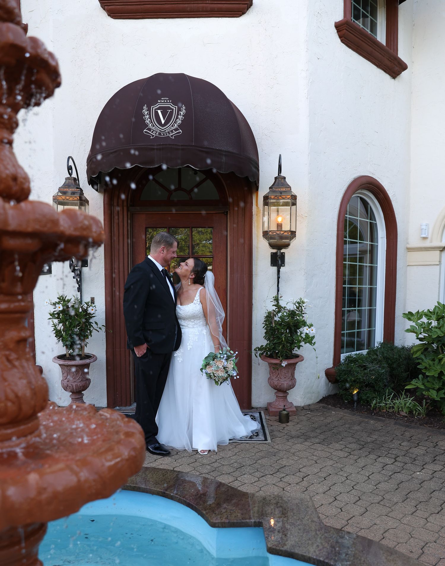 Bride and groom share a romantic moment near a fountain outside an elegant white venue with a brown awning.