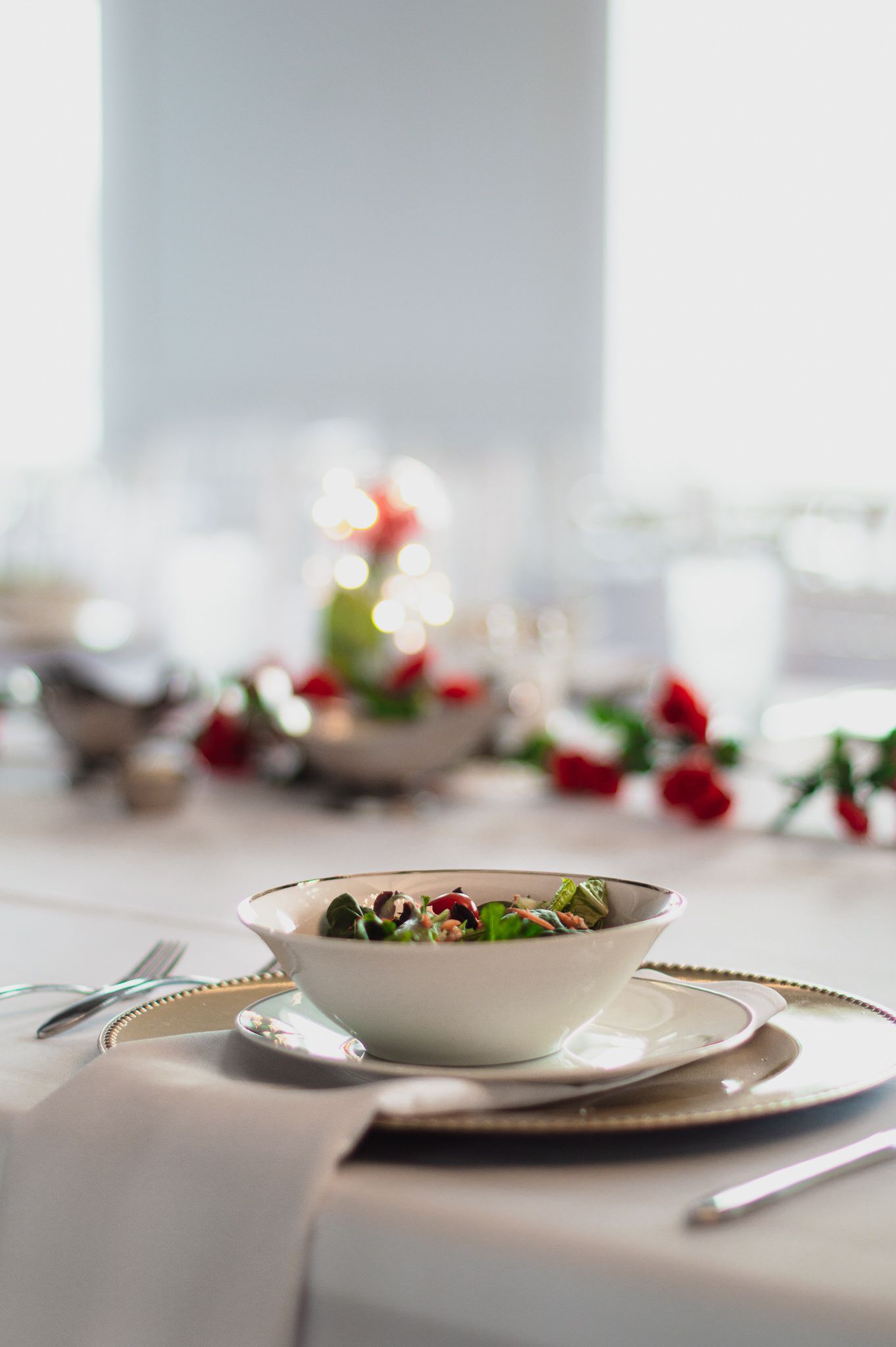 Elegant table setting with a white bowl on gold-rimmed plate, red roses, and candlelight in the background.