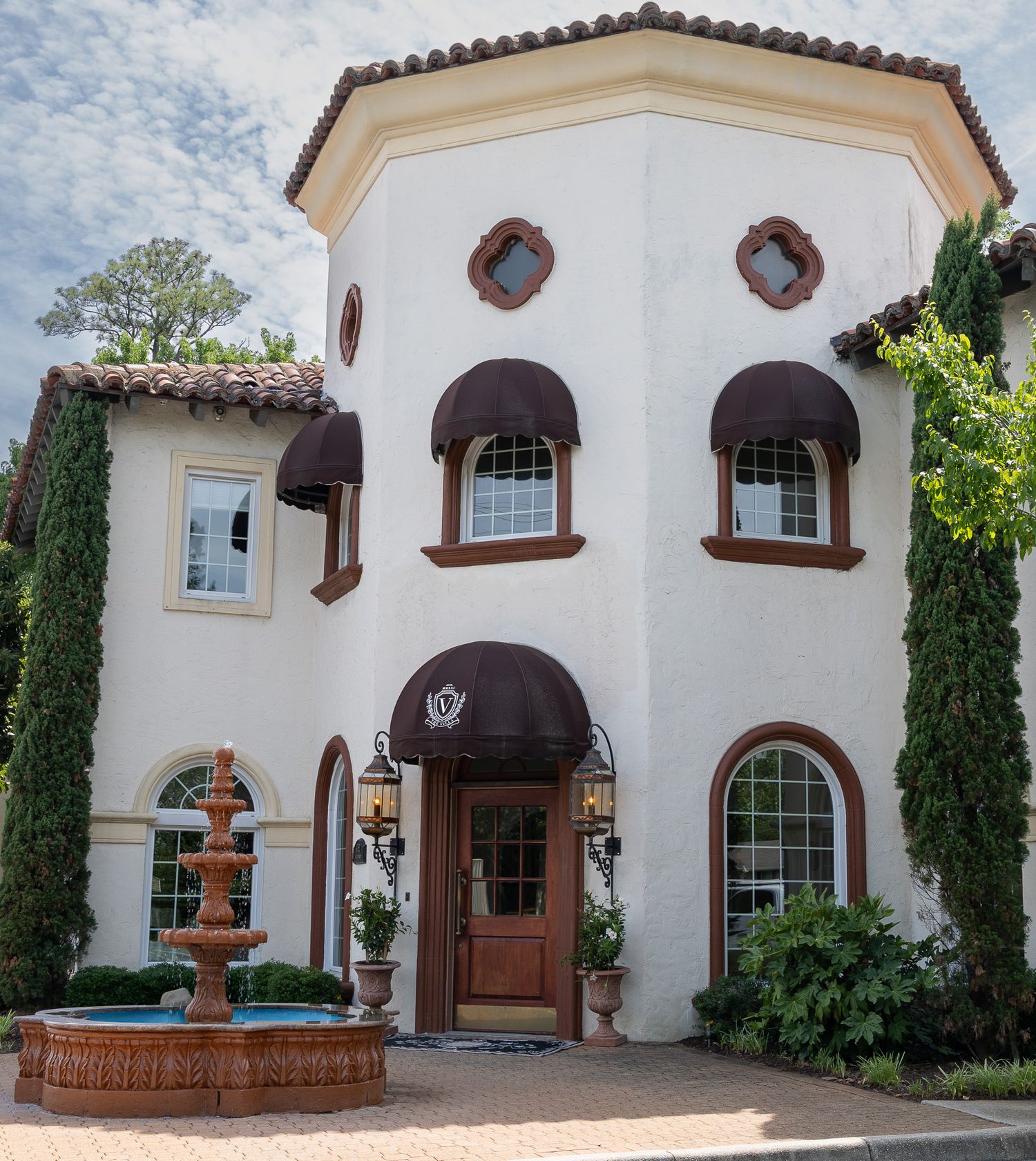 Spanish-style white stucco home with terracotta roof, arched windows, brown awnings, and a decorative fountain in the front.