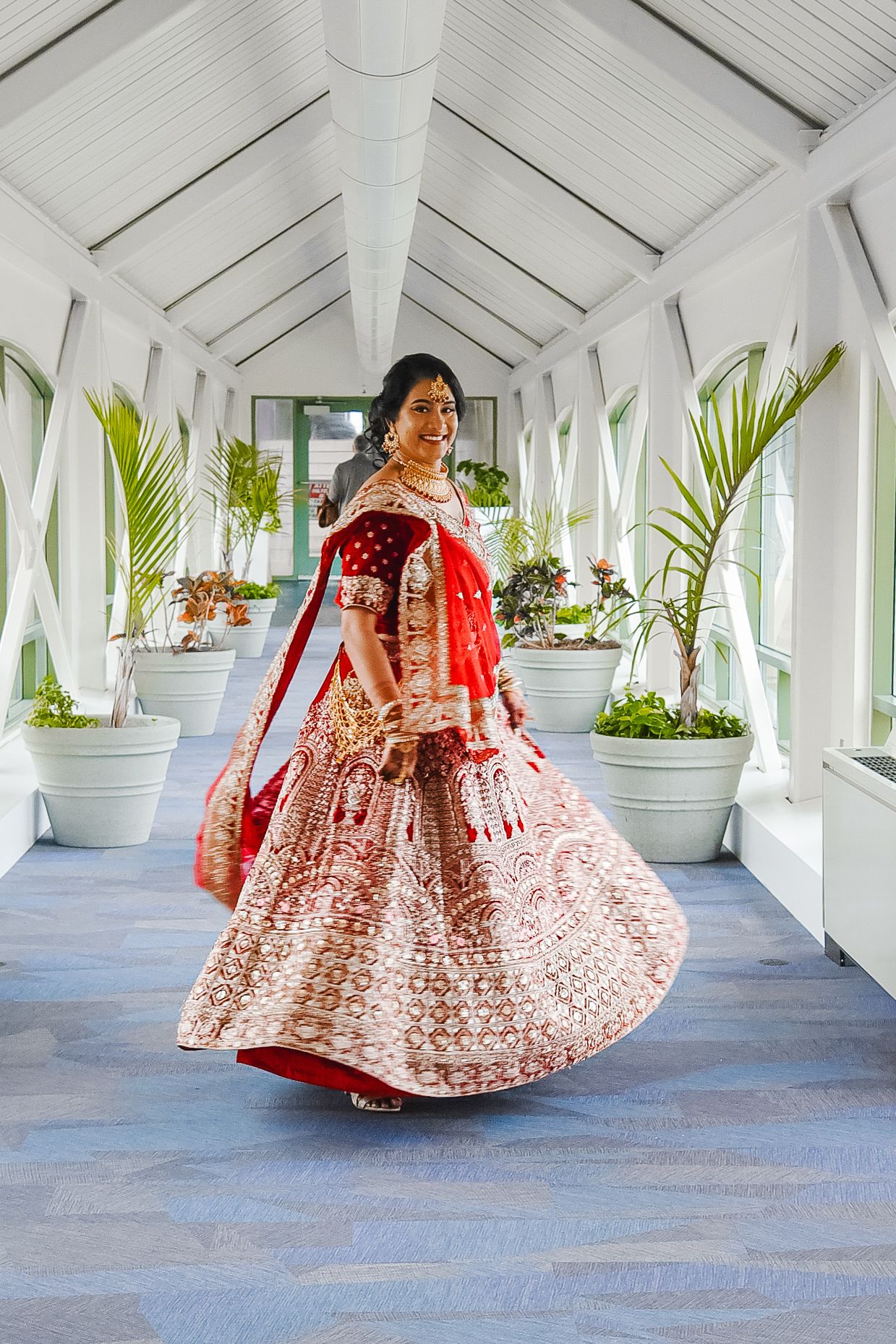 Bride in red and cream lehenga twirling in white hallway with tropical plants.