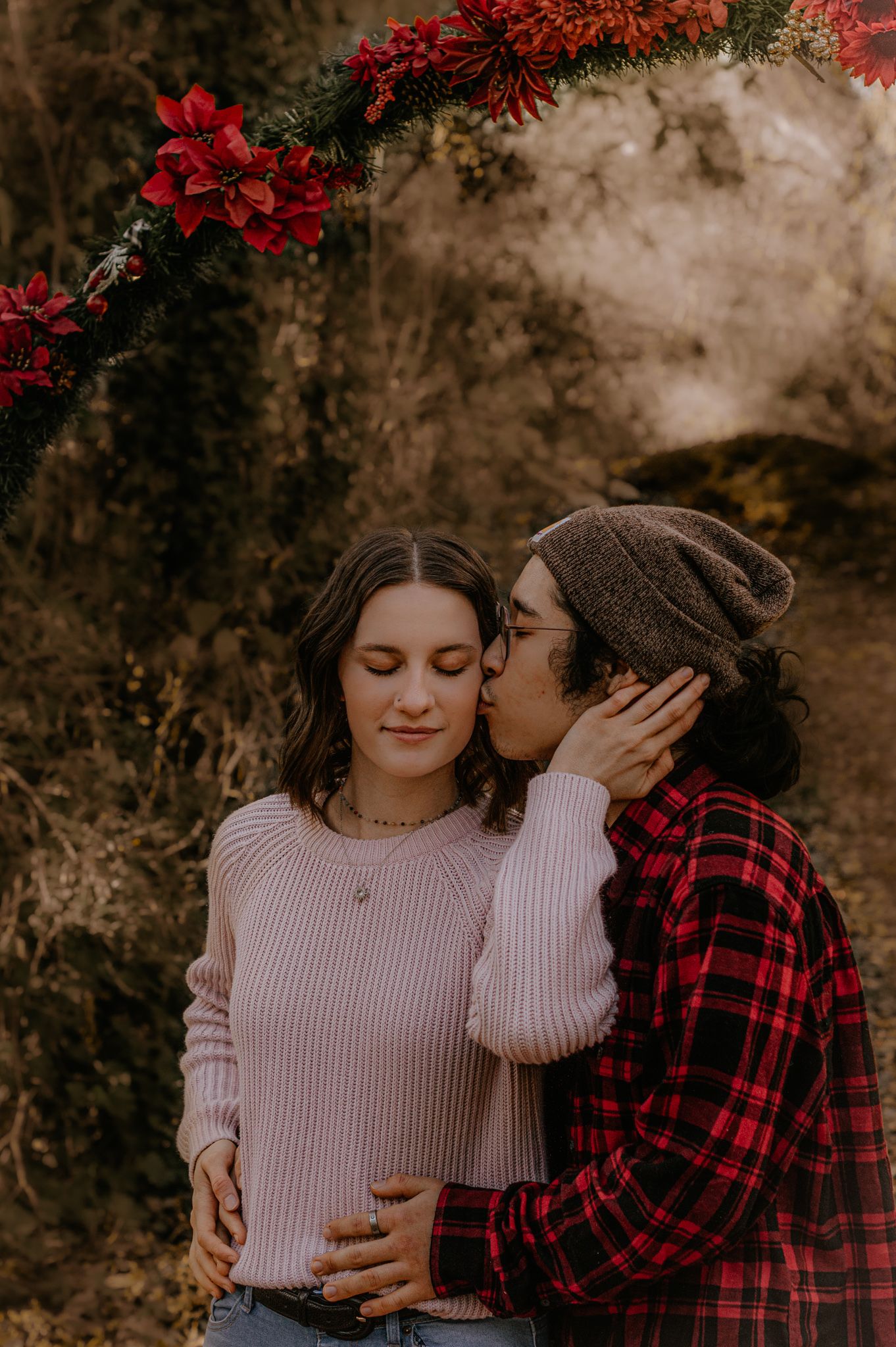 Couple sharing intimate moment among red flowers against rustic wall.