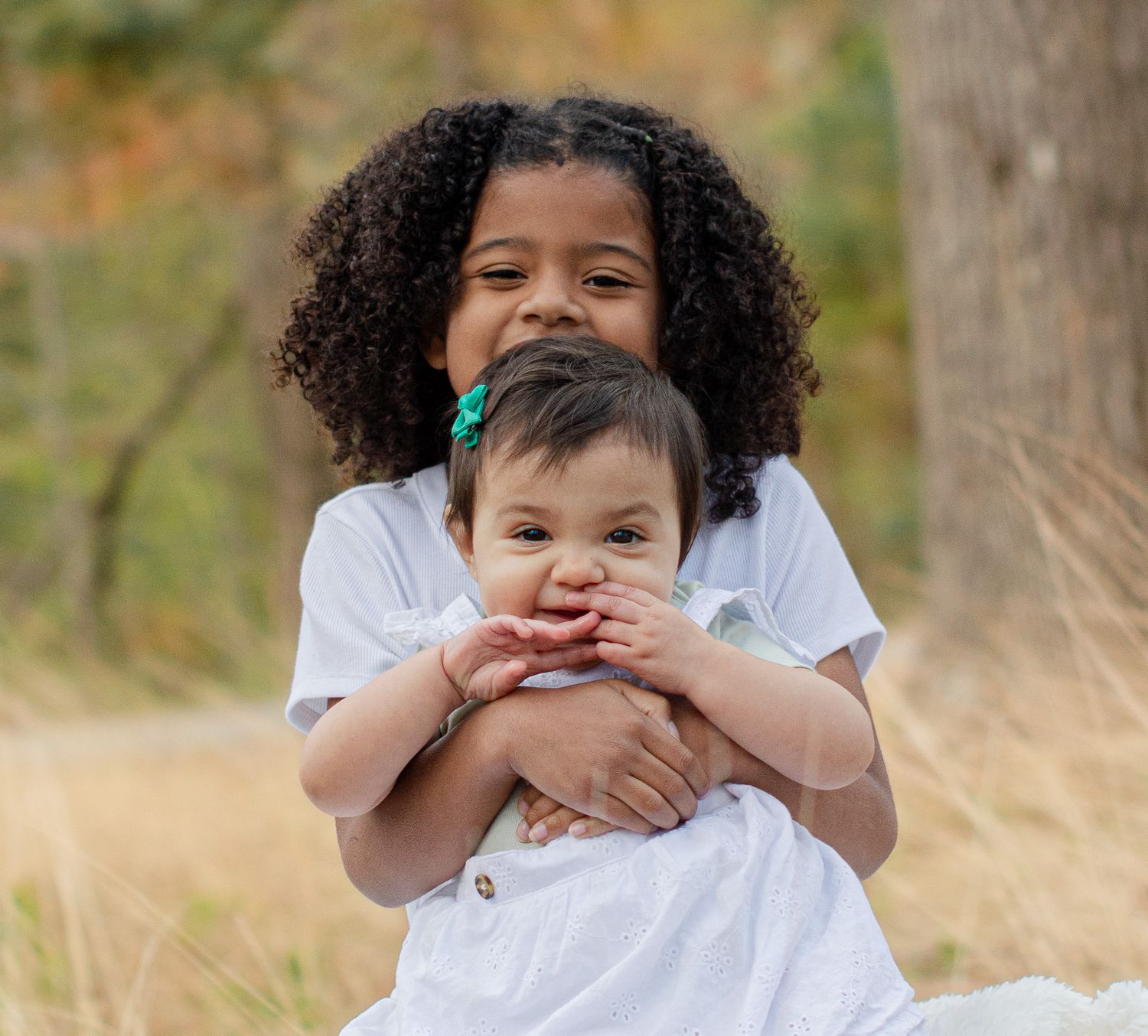 Two young sisters in white dresses pose outdoors, older girl hugging baby sibling in a golden field.