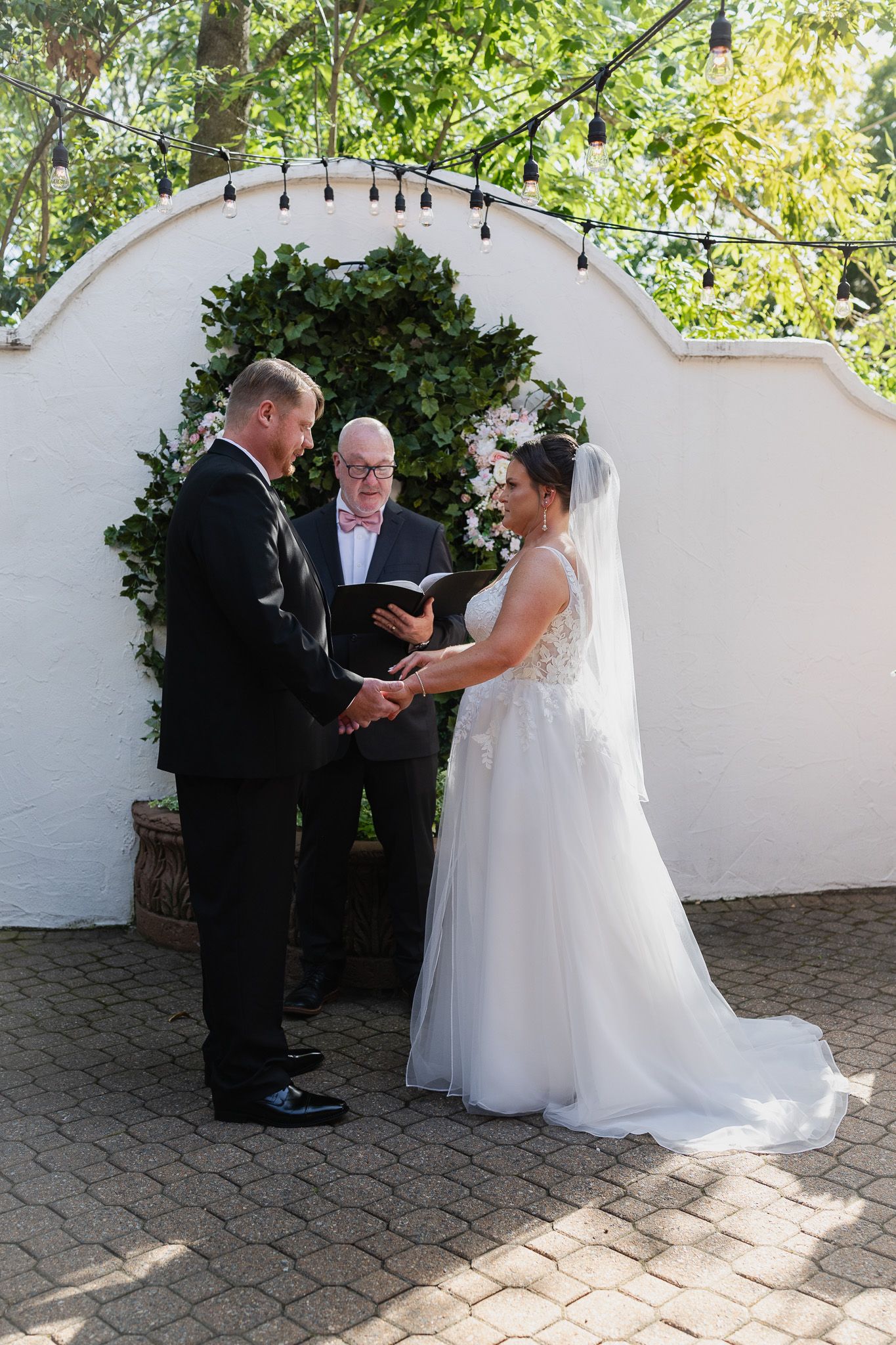 Bride and groom hold hands during outdoor wedding ceremony before officiant at ivy-covered arch.