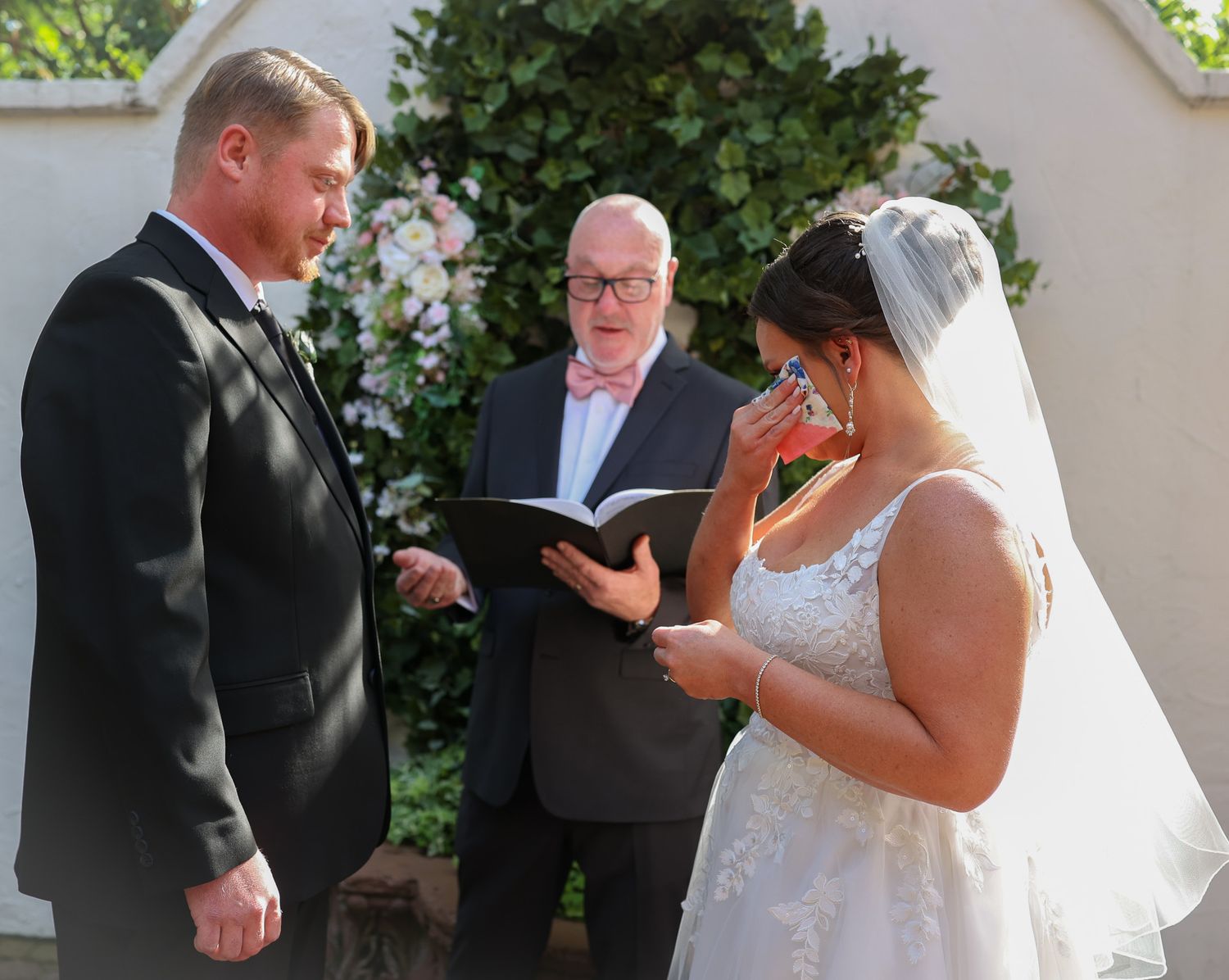 Bride wiping tears during outdoor wedding ceremony while groom and officiant stand before floral arch.
