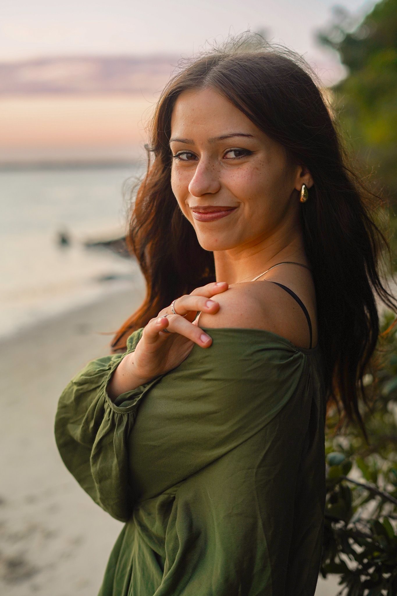 A person in an olive green sweater poses on a beach at sunset with warm golden light in the background.
