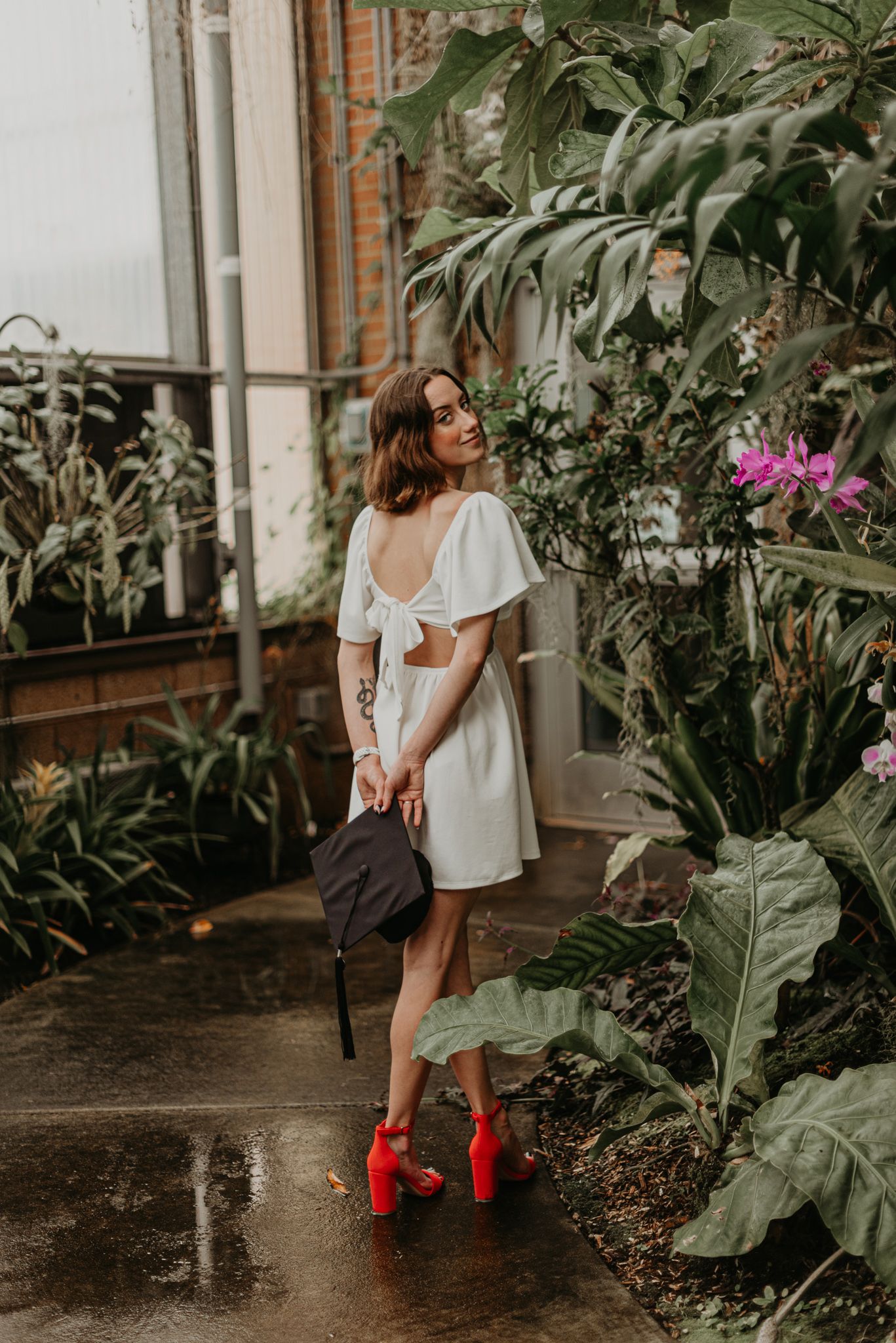 A person in a white dress and bright red boots stands in a lush greenhouse setting with tropical plants.