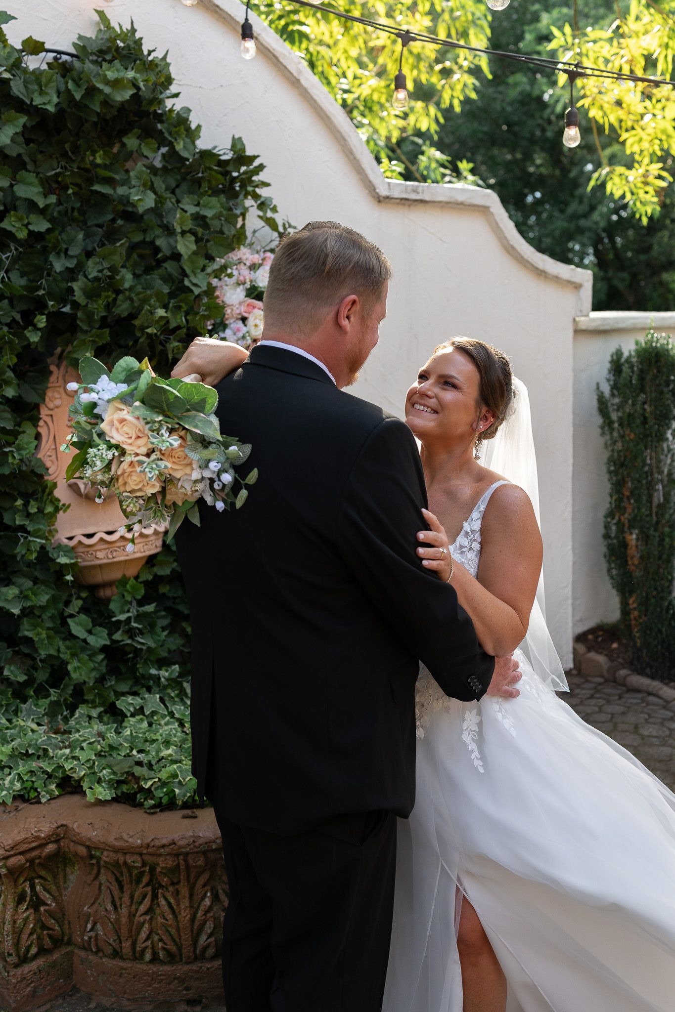 Bride in white gown smiles at groom in black suit holding bouquet outdoors at elegant garden wedding venue.