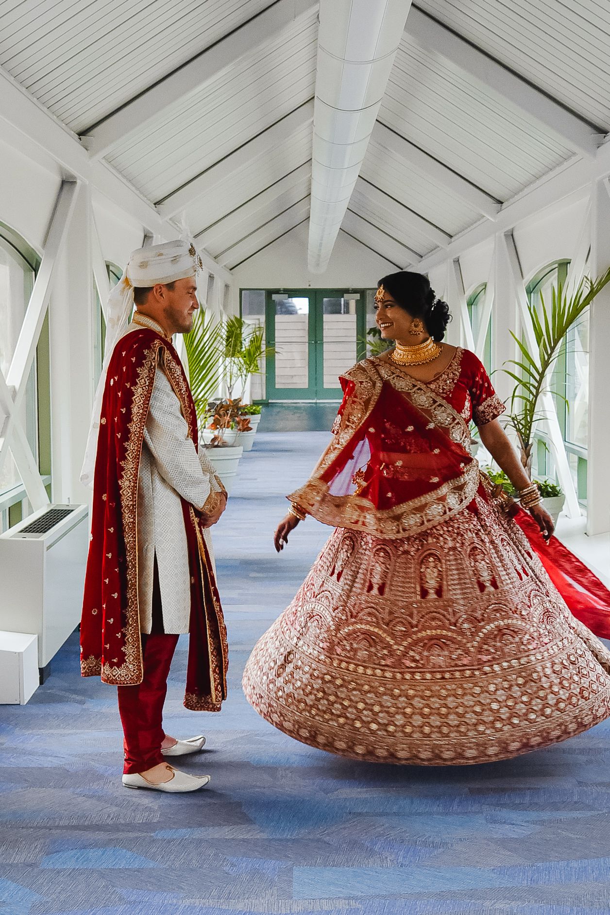Traditional Indian wedding couple in red and pink lehenga photographed in white corridor.