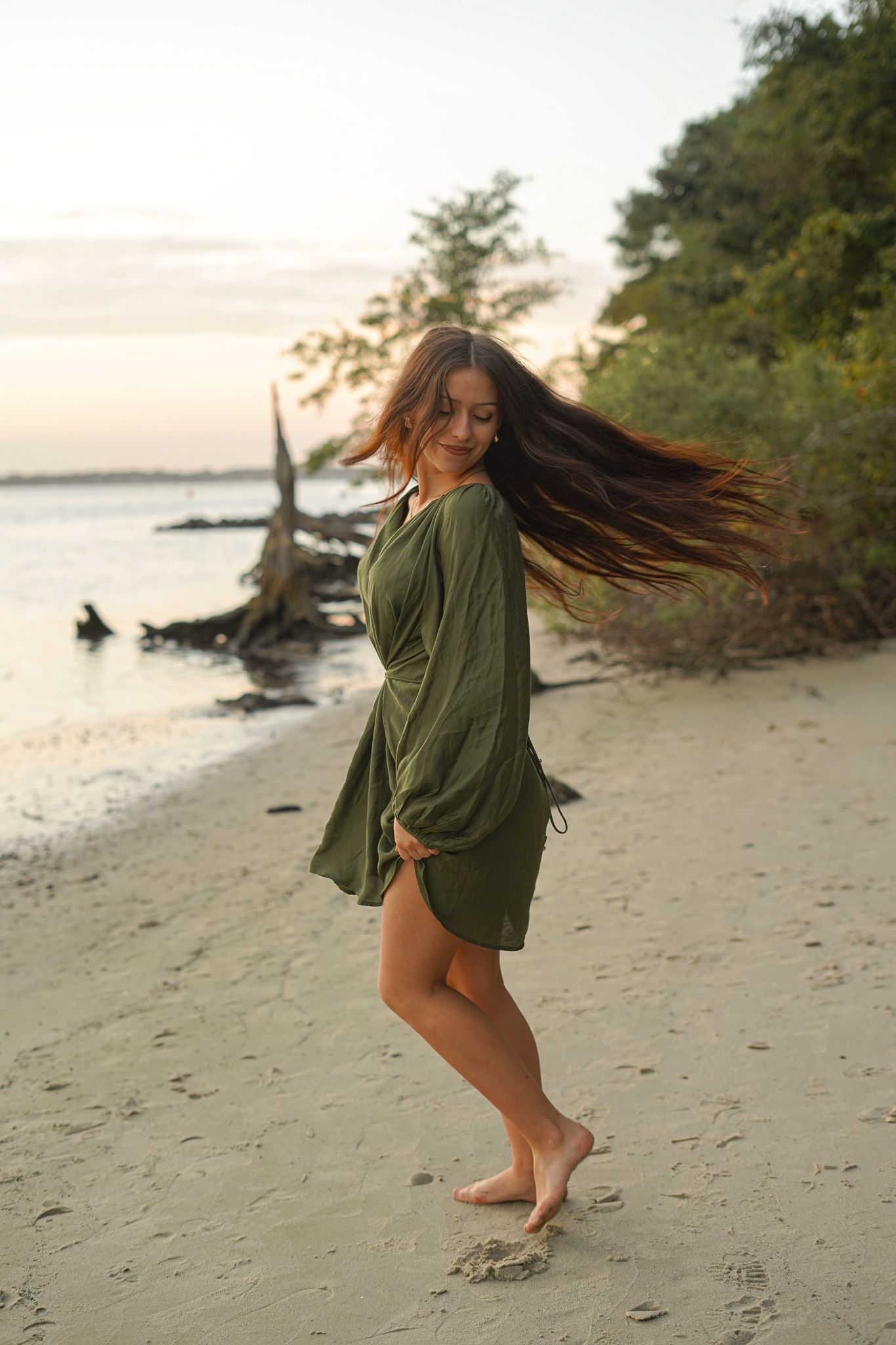 A person in an olive green dress dances and twirls joyfully on a sandy beach at sunset with ocean waves in the background.