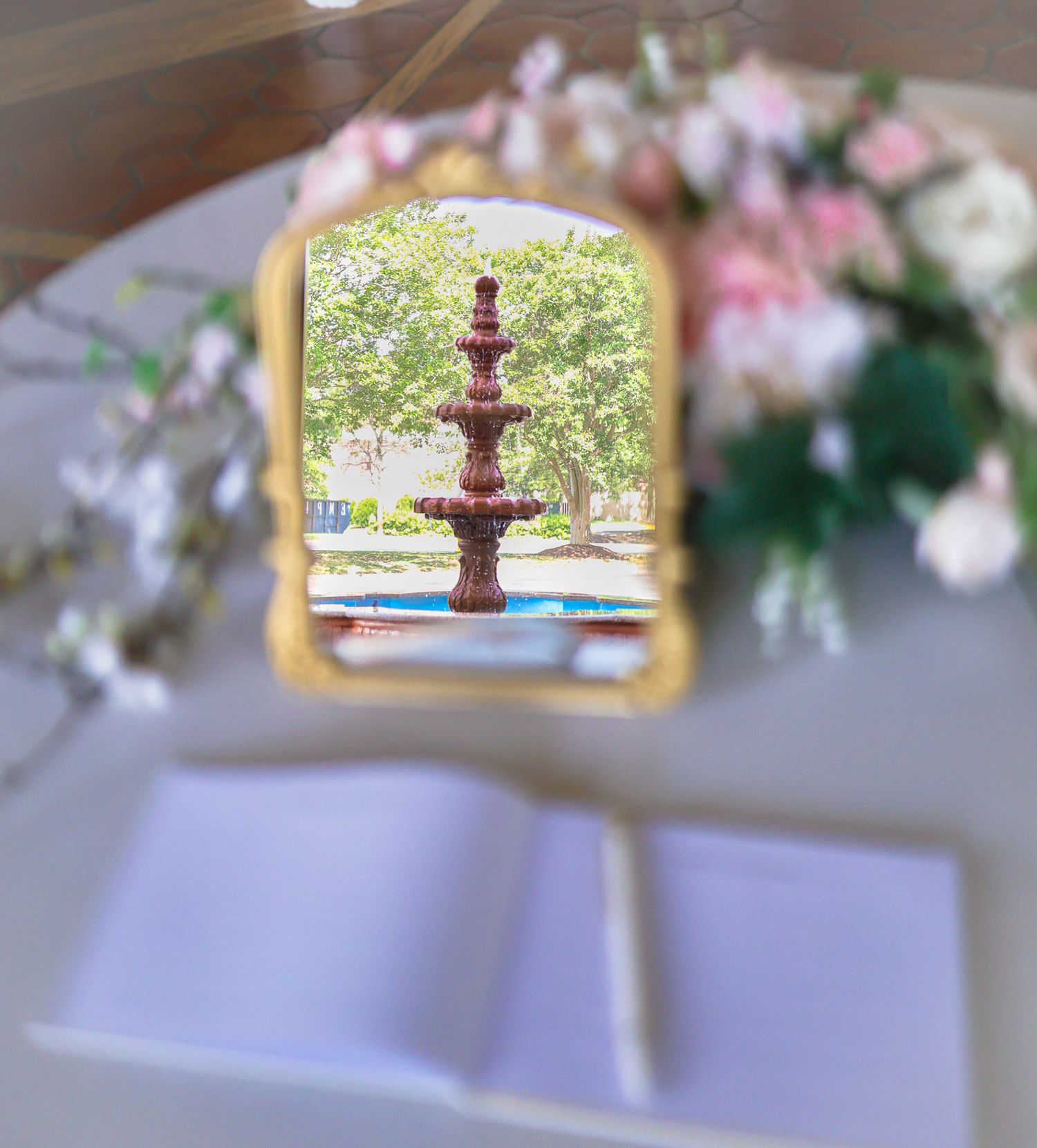Elegant wedding sign-in table with open guest book, gold mirror reflecting outdoor fountain, and pink floral arrangements.