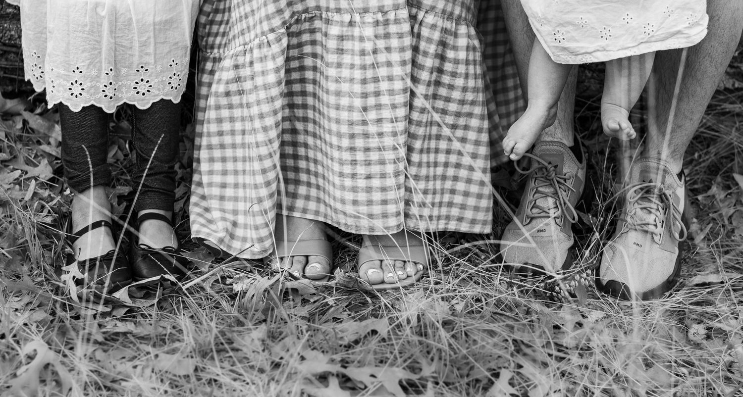 Black and white photo of children standing barefoot and in shoes on grass, showing only their legs and feet.