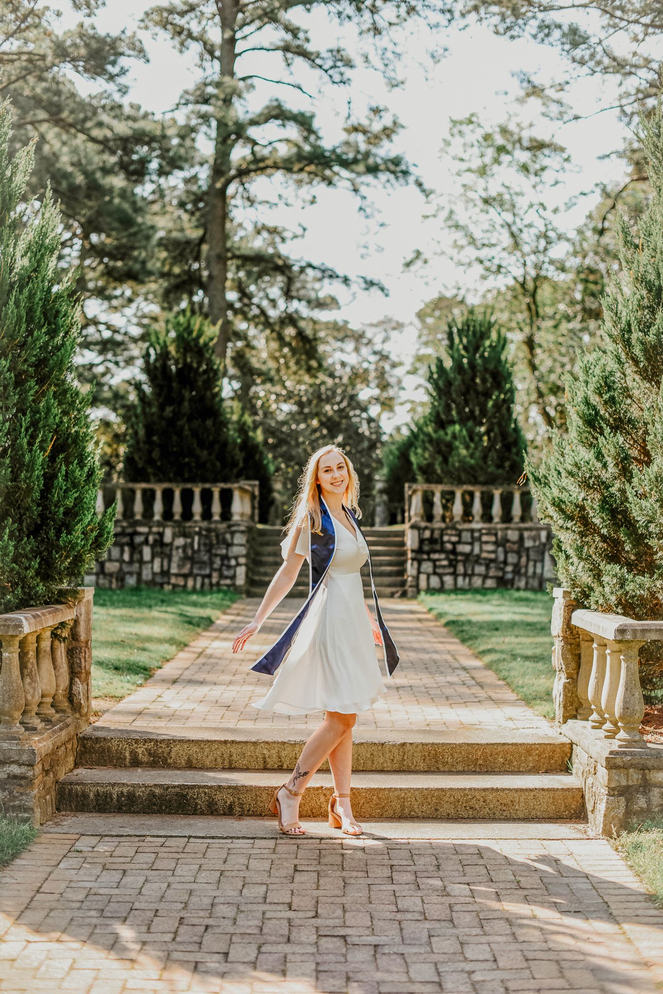 Graduate in white dress poses on stone steps with decorative pillars and evergreens.