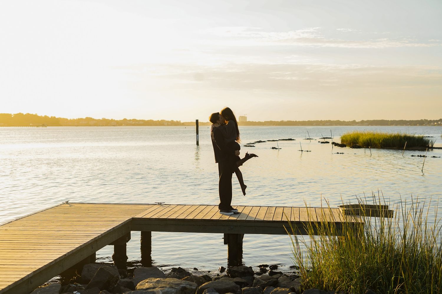 A wooden boardwalk extends over a peaceful coastal marsh at sunset with golden light reflecting on calm waters.
