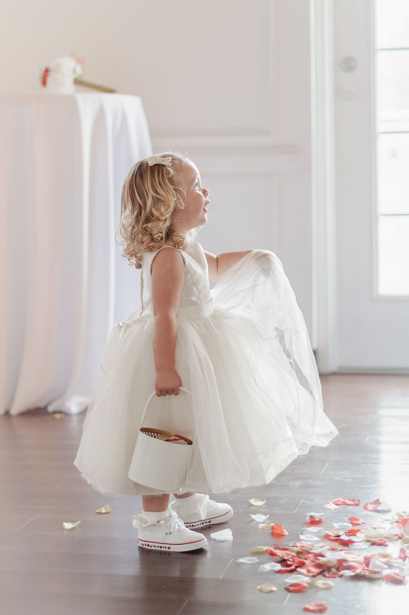 Adorable flower girl in white dress and sneakers holding a basket with rose petals scattered on the floor at a wedding.