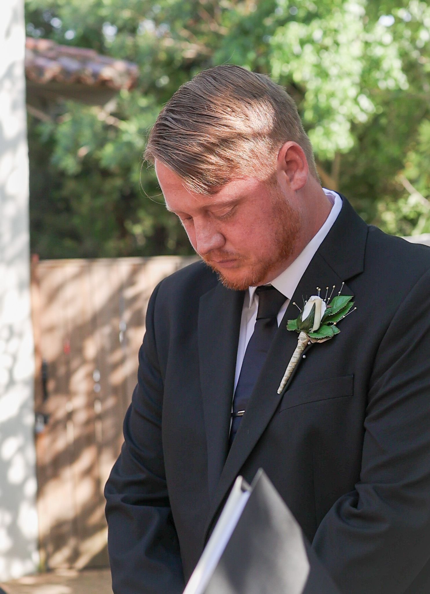 Groom in dark suit with boutonniere reads vows during outdoor wedding ceremony.