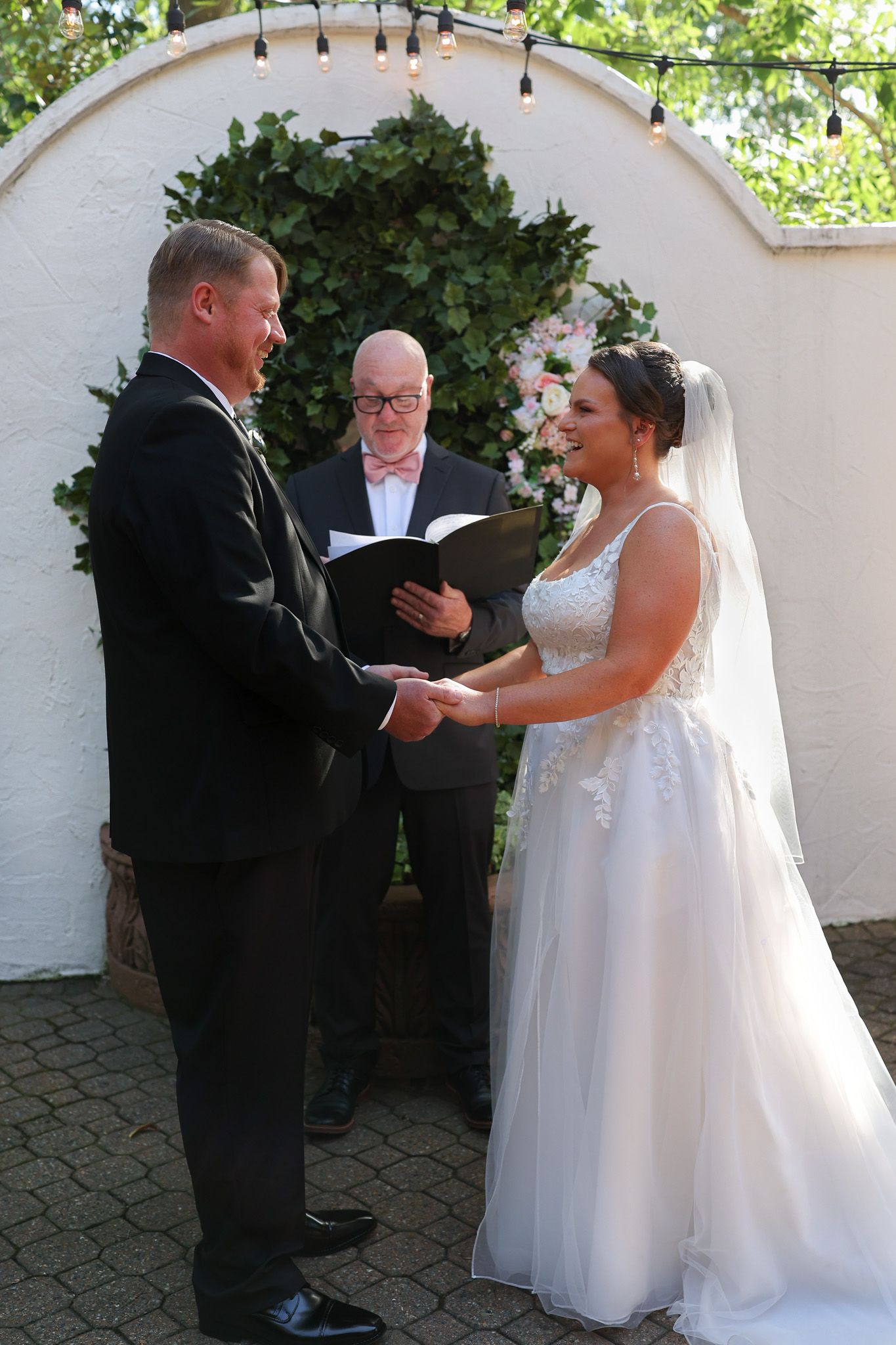 Bride and groom exchange vows at outdoor wedding ceremony with officiant holding book before floral arch.