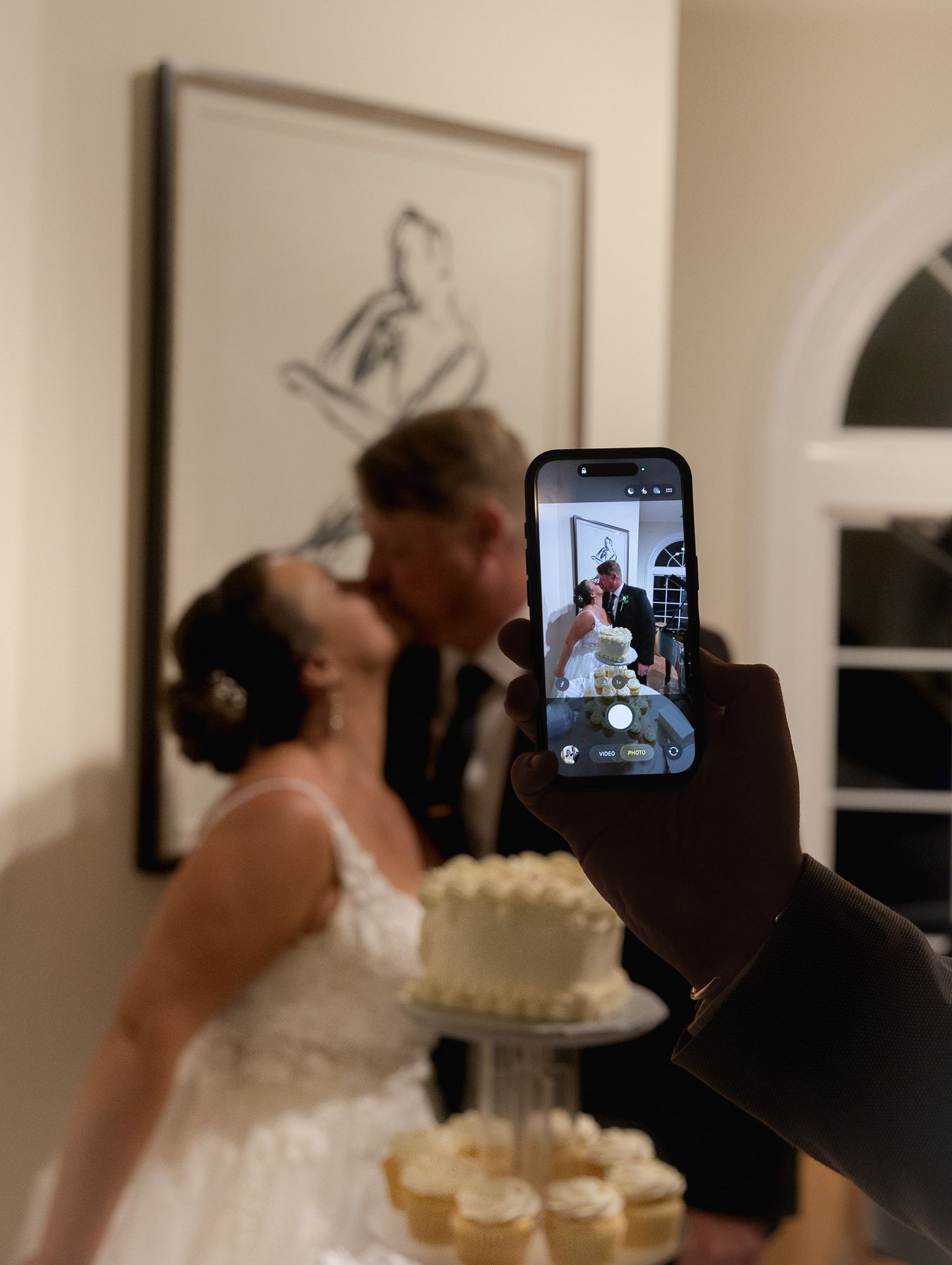 Guest photographs bride and groom kissing behind wedding cake with smartphone at reception.