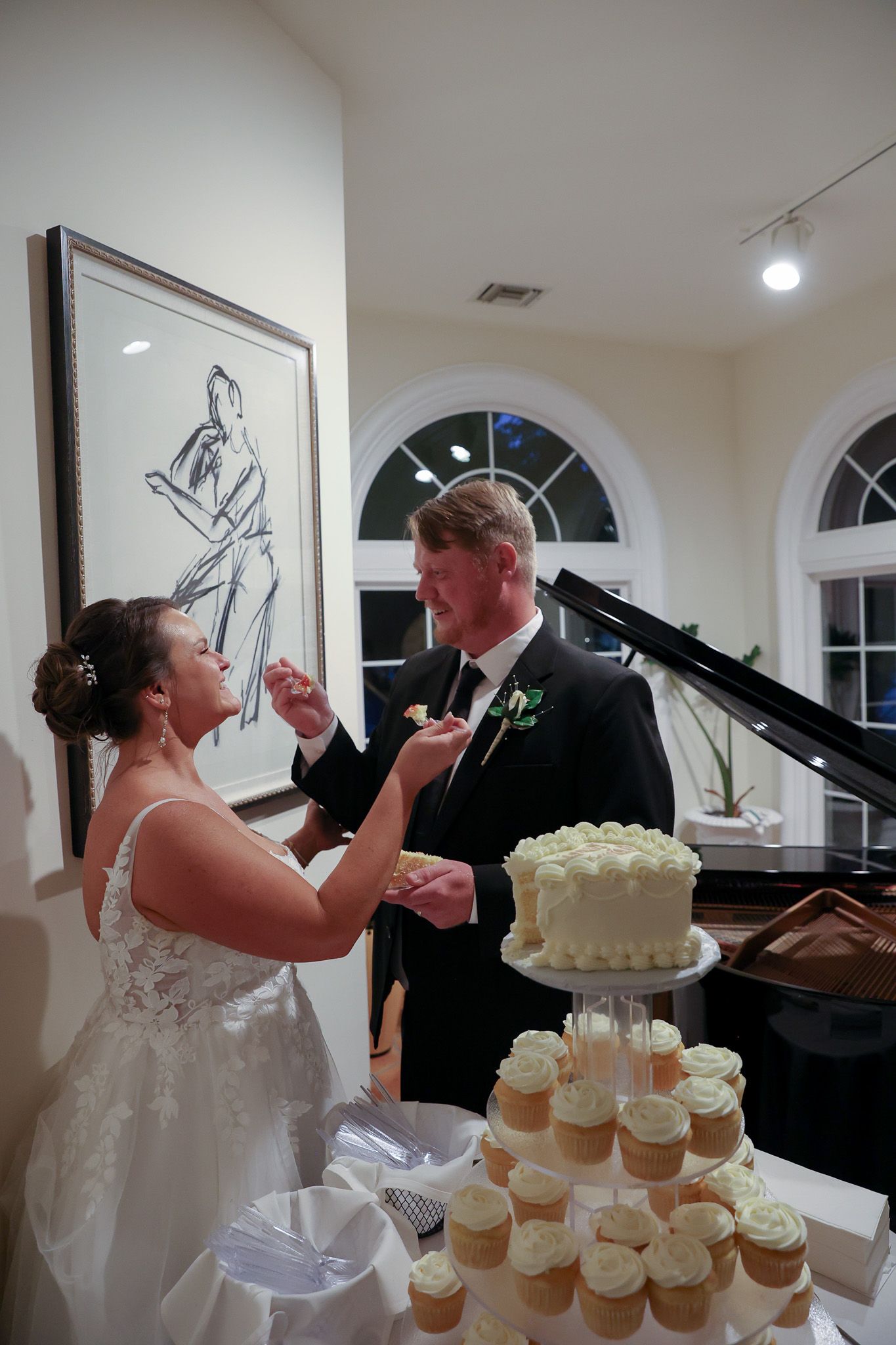 Bride and groom feeding each other wedding cake at reception, with white tiered cupcake tower and grand piano in background.
