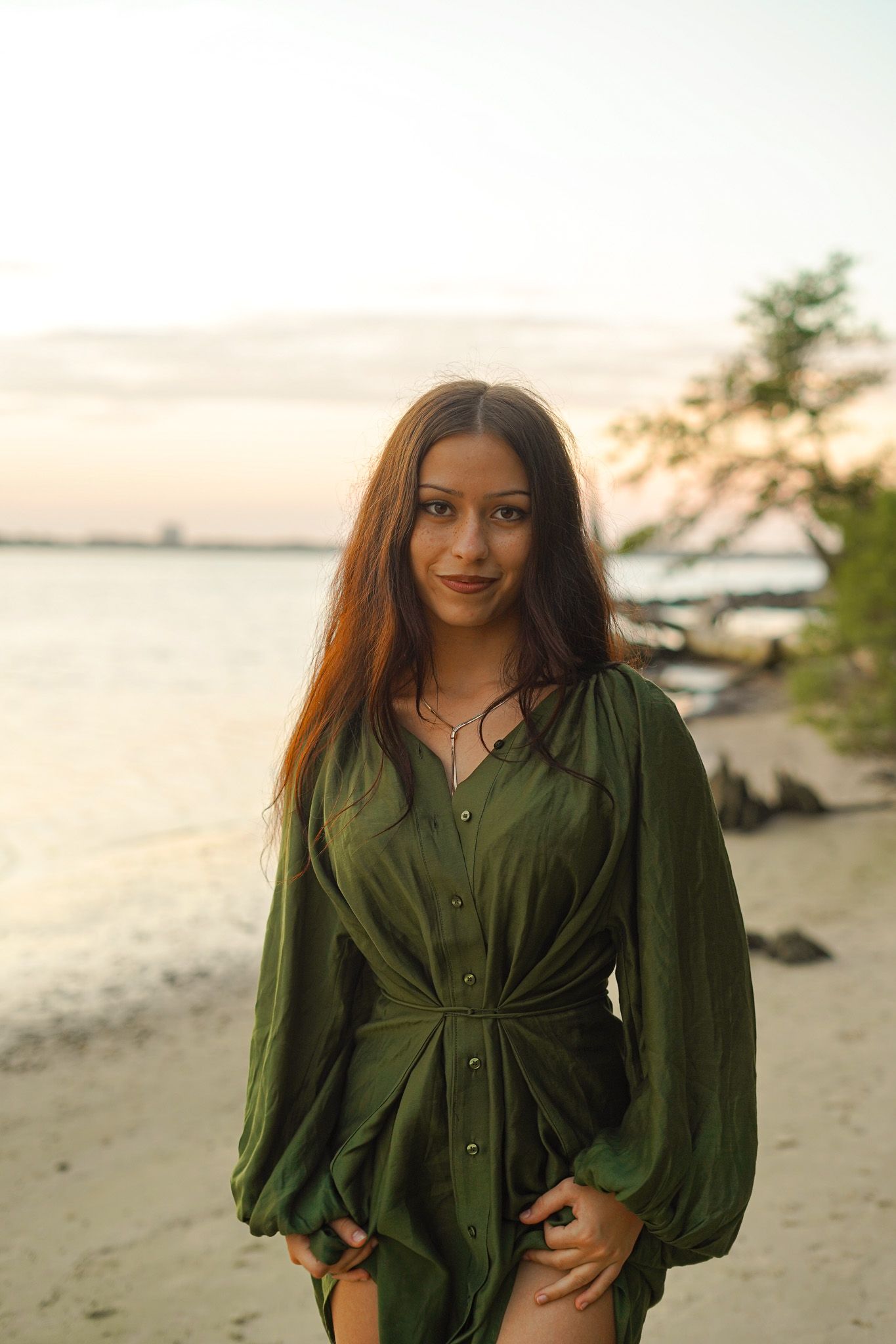 Woman in green button-down dress poses on beach at sunset.