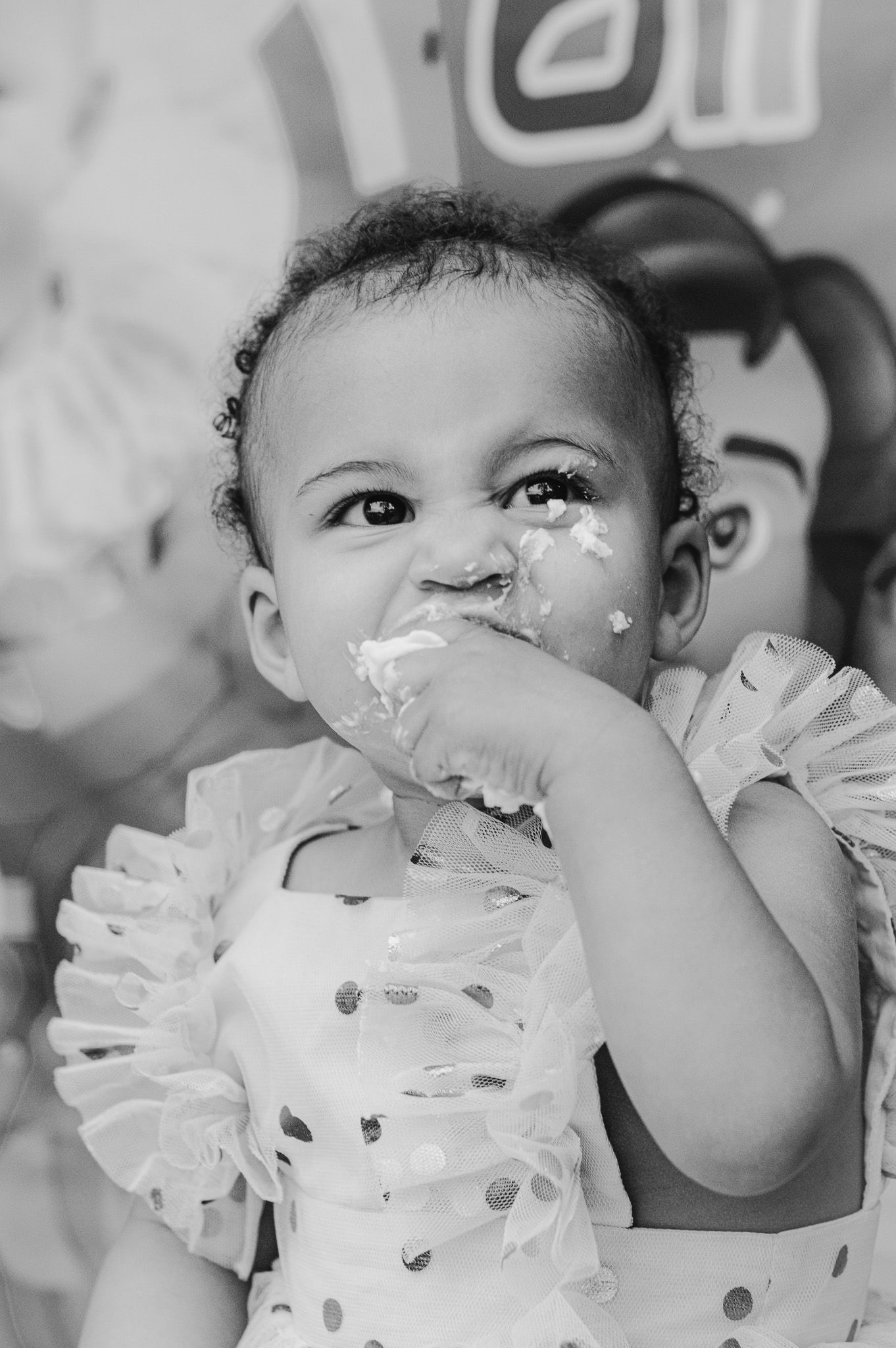 Baby wearing polka dot outfit with cake frosting on face during first birthday celebration.