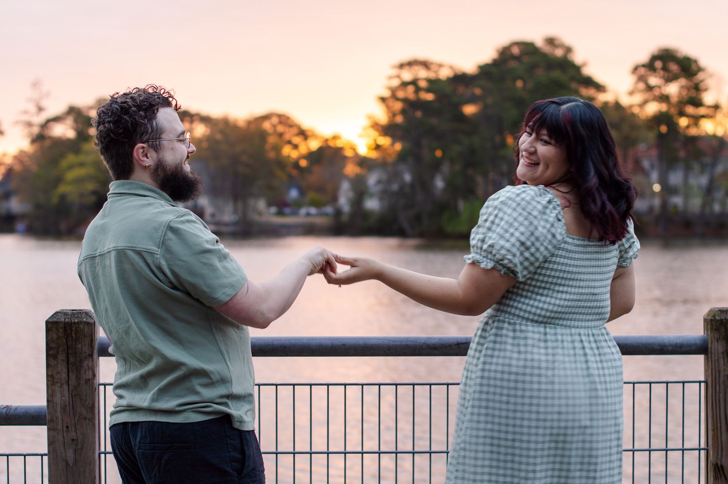 Couple holding hands and smiling at sunset by a lake, man in green shirt, woman in gingham dress.