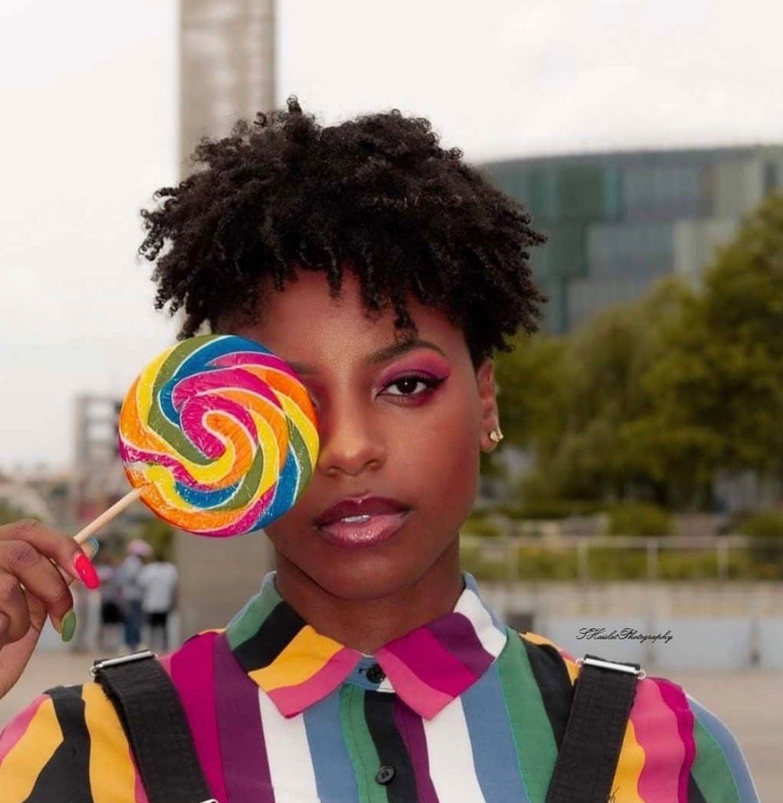 A person with curly hair holds a colorful swirled lollipop while wearing a striped multicolored outfit.