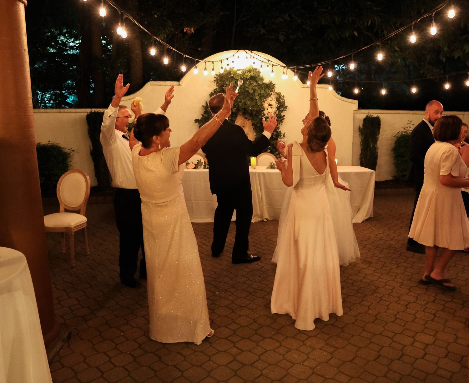 Guests in white gowns dance at an outdoor evening wedding reception with string lights and floral arch.